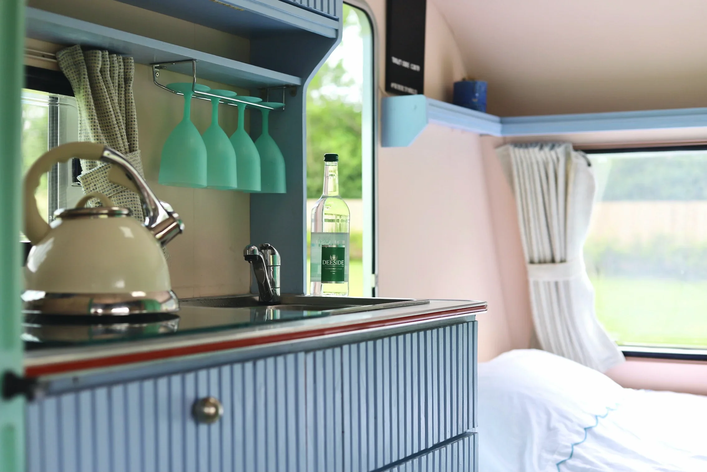 Interior of a vintage blue camper with a small kitchen area featuring a sink, a kettle, and hanging turquoise glasses. There are windows with striped curtains and a bed with white bedding visible at the back.
