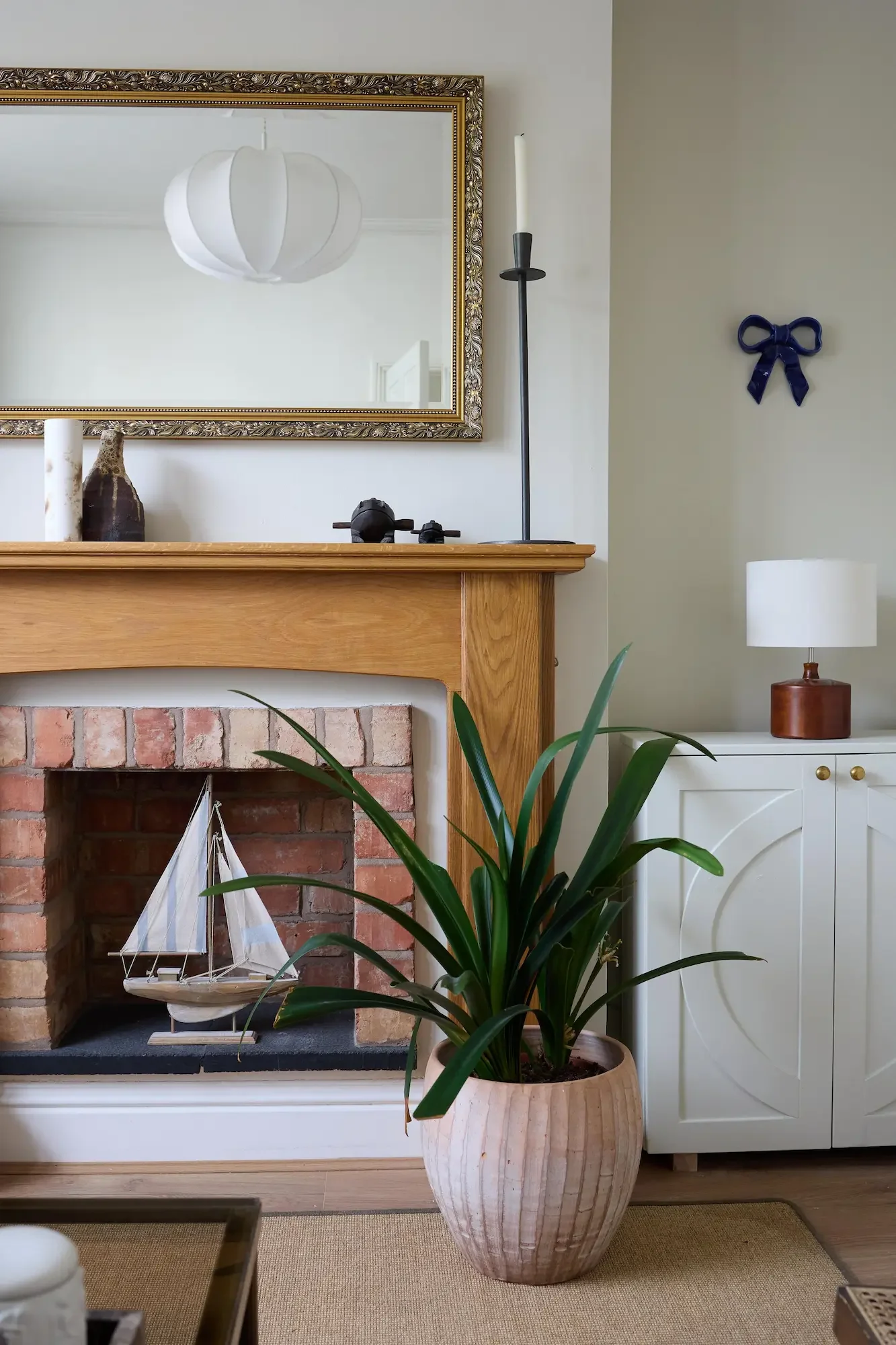 Living room with a fireplace, large potted plant in front, mirror above the fireplace, ship model inside the fireplace, candle holder and lamp on the mantel, and a white cabinet with a lamp on top to the side.
