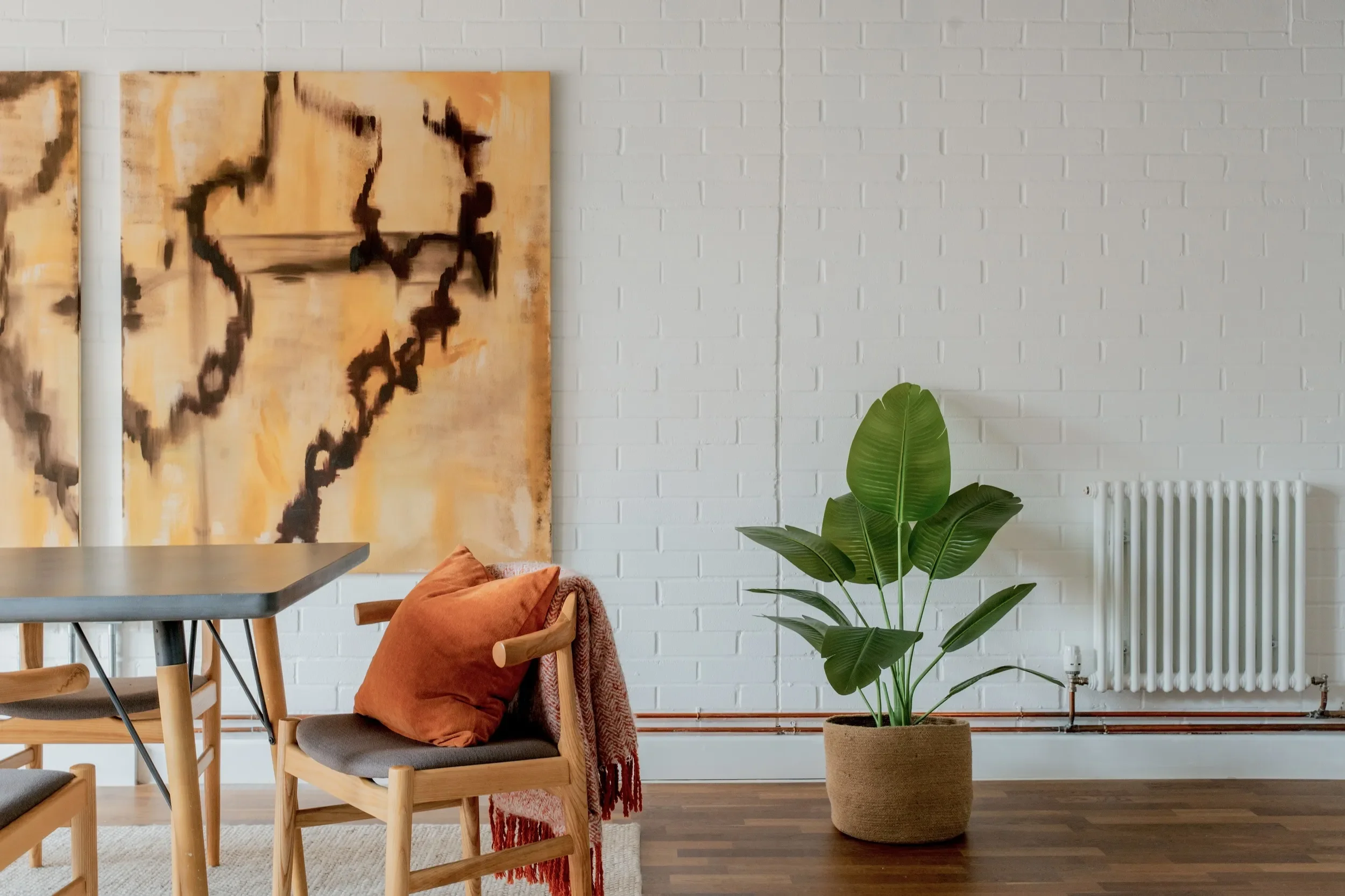 A modern interior space with a white brick wall, wooden furniture including a table and chairs, a large potted green plant, and abstract artwork hanging on the wall.