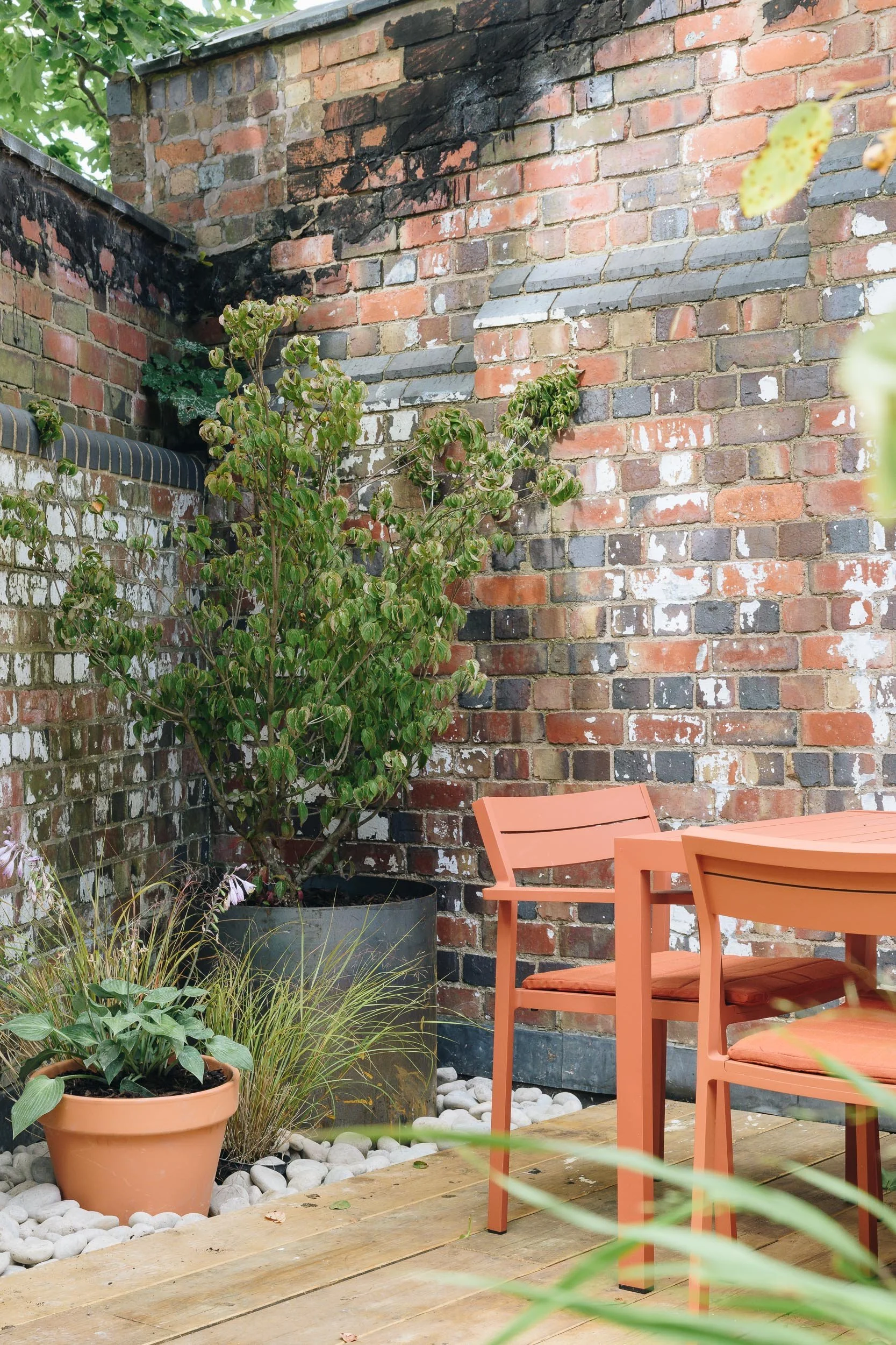 Outdoor patio area with orange chairs around a matching table, potted plants, a brick wall with some black soot marks, and green foliage.