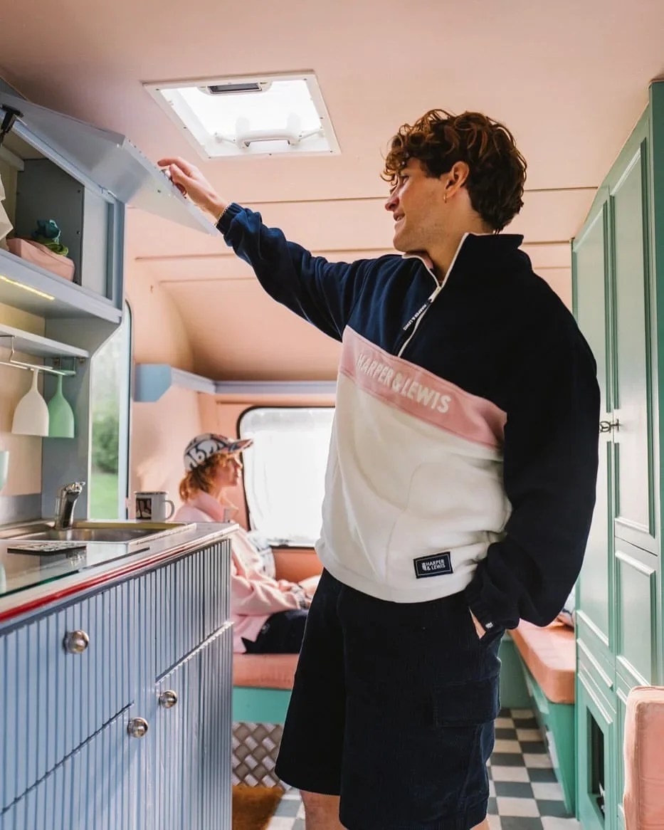 A young man with curly hair reaching into an overhead cabinet inside a retro-patterned camper with pastel green cabinets, a checkered floor, and a seating area where a young woman wearing a pink cap and sweatshirt sits by a window.