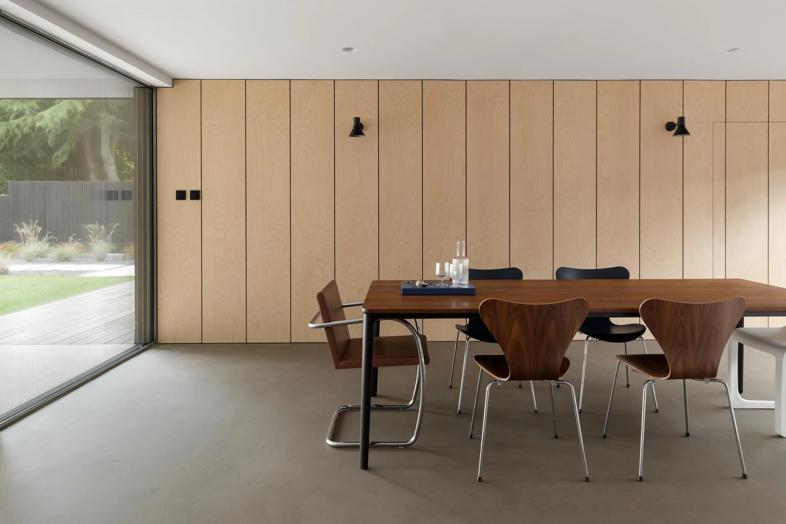 Modern dining room with wooden wall panels, a wooden dining table with black chairs, a tray with glasses, and large sliding glass door showing greenery outside.