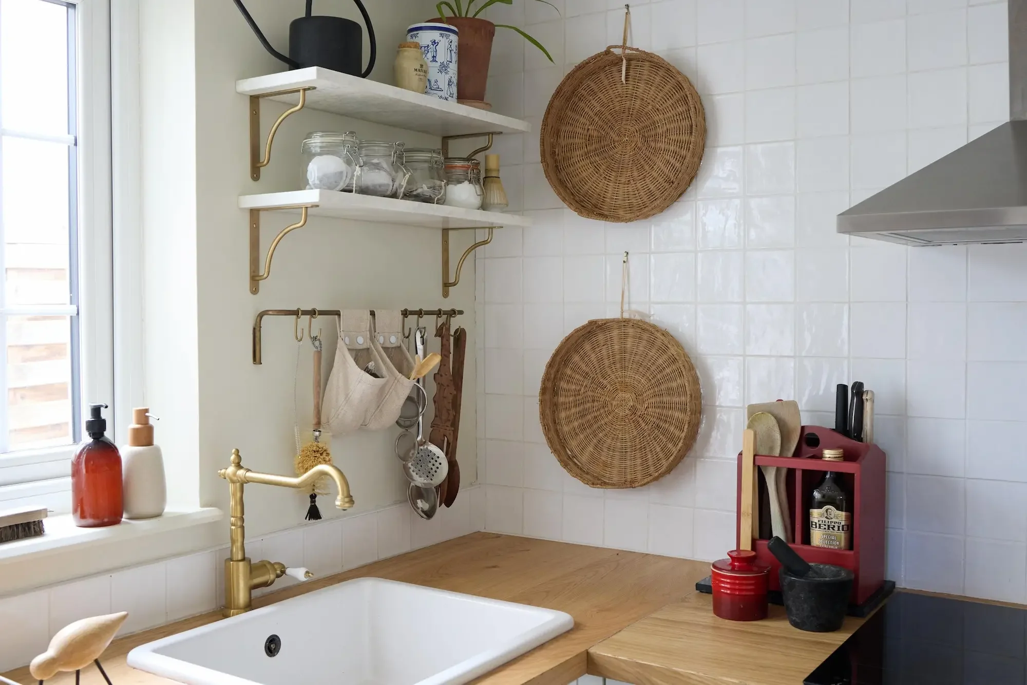 A kitchen sink island with a white farmhouse sink, brass faucet, and a wooden countertop. Above it, white shelves hold jars, plants, and decorative items. Two round woven baskets hang on a white tiled wall. The window to the left lets in natural light. Various soap bottles, kitchen utensils, and black and red containers are on the countertop and a small black drying rack.