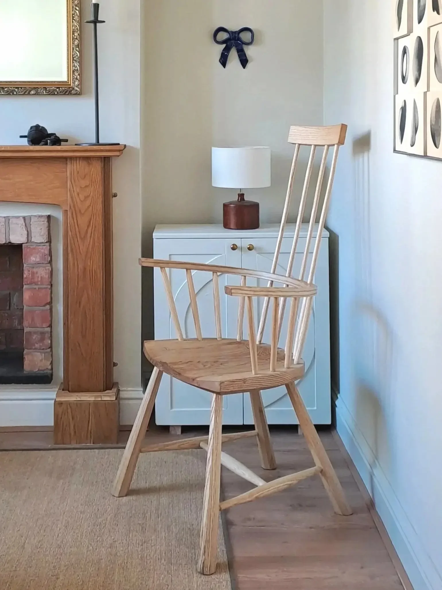 A wooden armchair with vertical slats on the backrest, placed on a light-colored carpet, in front of a small white cabinet with gold knobs, a table lamp with a white shade and wooden base, a brick fireplace with a wooden mantel, and wall art including a black ribbon decoration and a framed art piece.