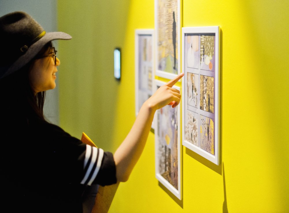 A woman wearing glasses, a hat, and a black shirt with white stripes on the sleeves points at an art display on a yellow wall in an art gallery.
