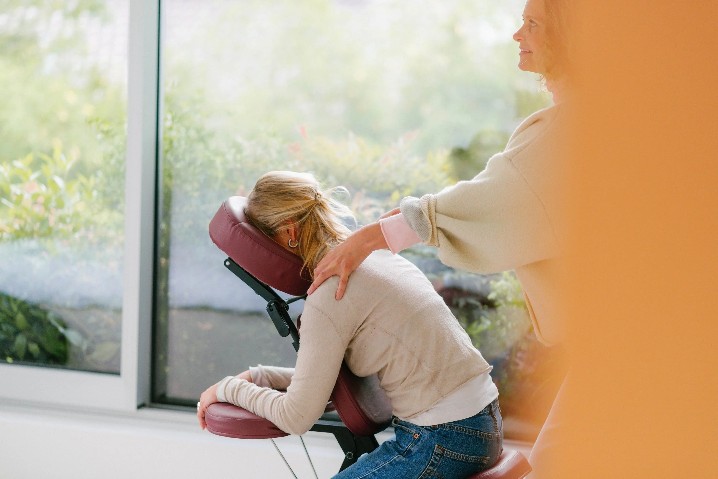 A woman providing a neck massage to a man seated in a massage chair in an indoor setting, with an office and reception area in the background.