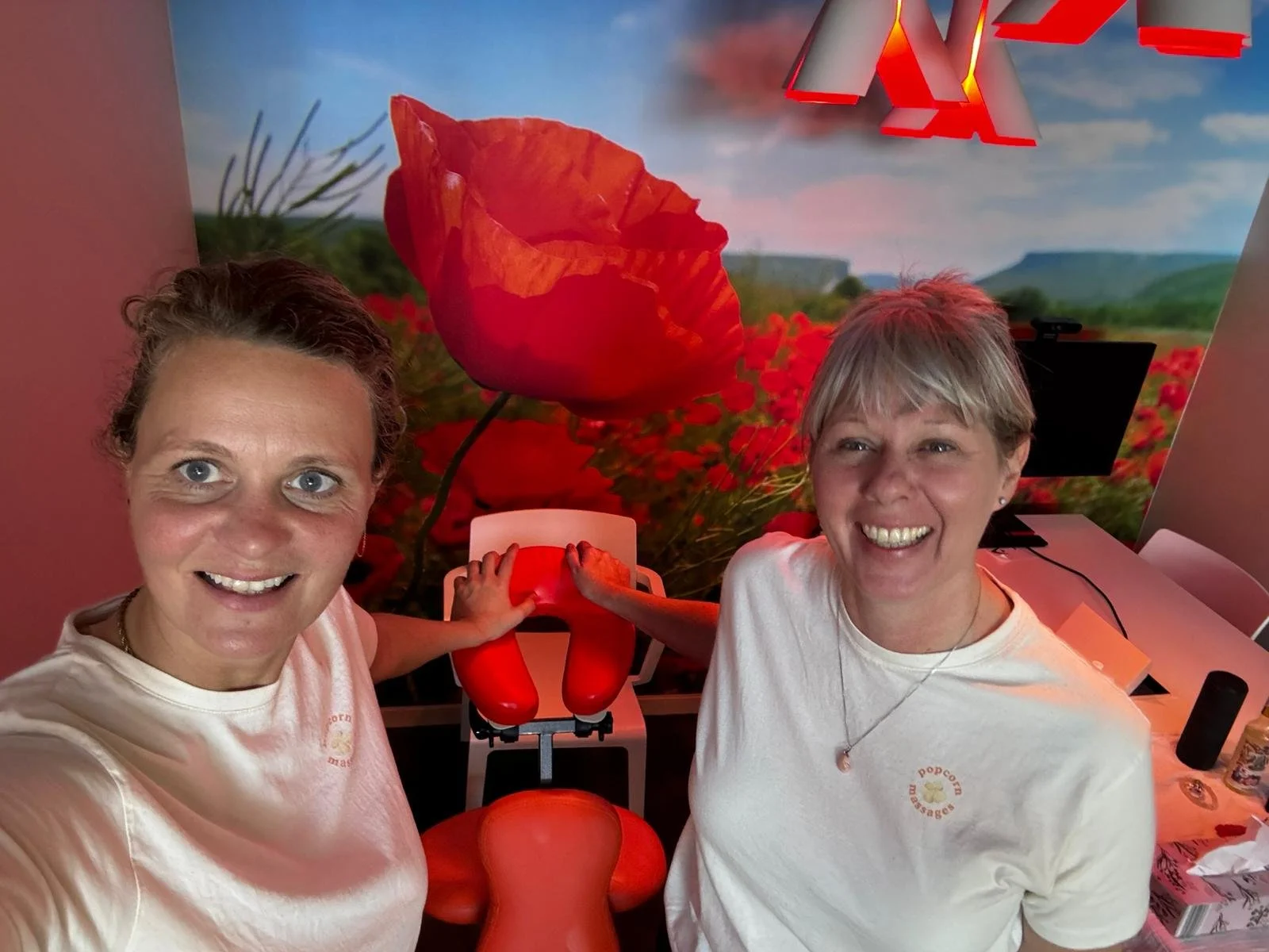 Two women smiling and touching a large red silicone vibrator in a room with a vibrant floral mural of red poppies.