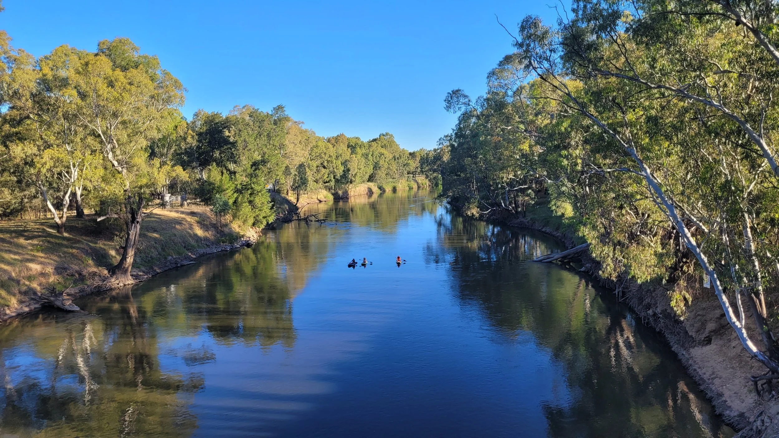 A calm river flowing through a lush green landscape with trees on both sides, with a few kayakers paddling on the water under a clear blue sky.