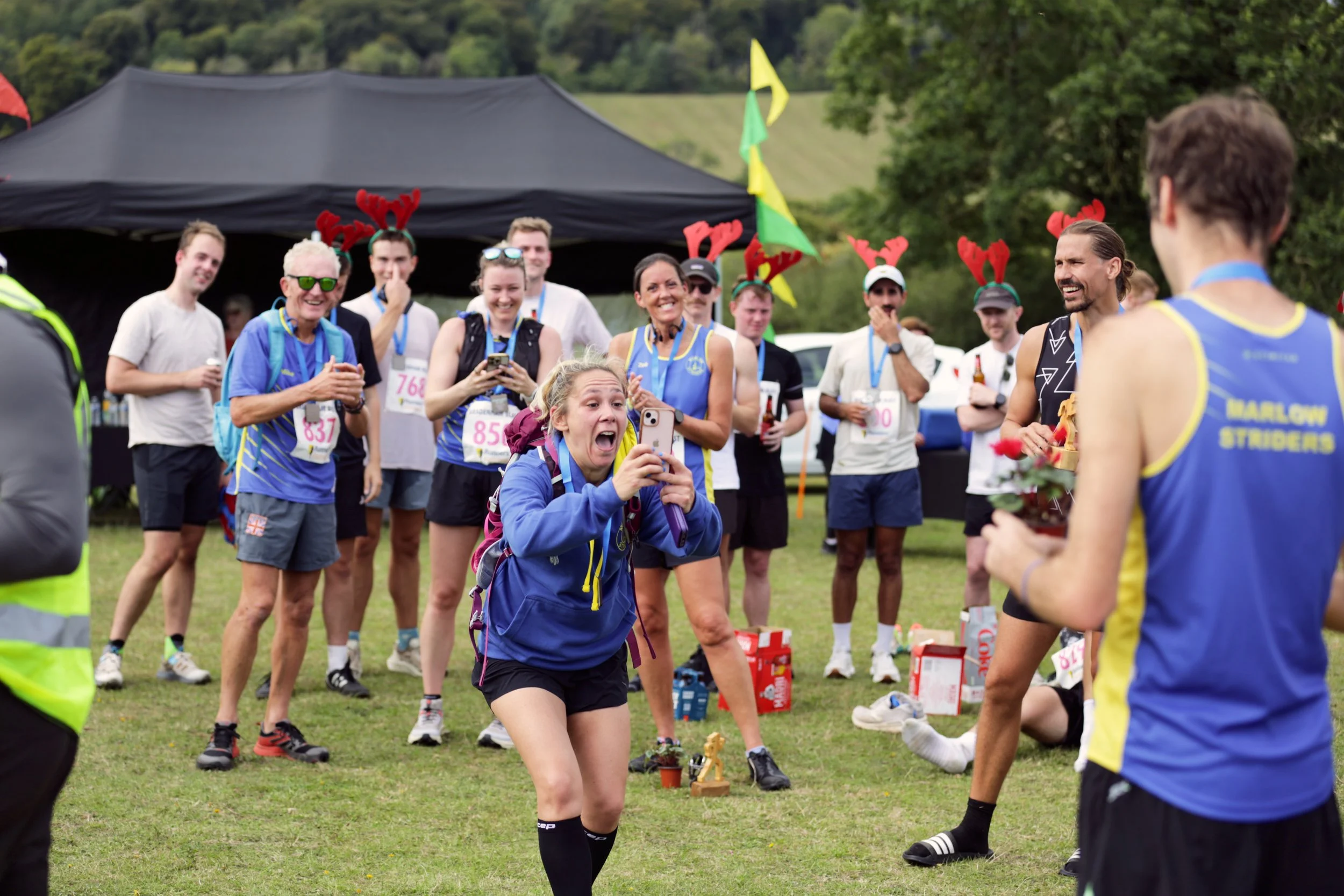Group of people at a sports event, some wearing reindeer antler headbands, with a woman in the foreground holding a phone and appearing surprised and happy. Banner flags and a tent are visible in the background.