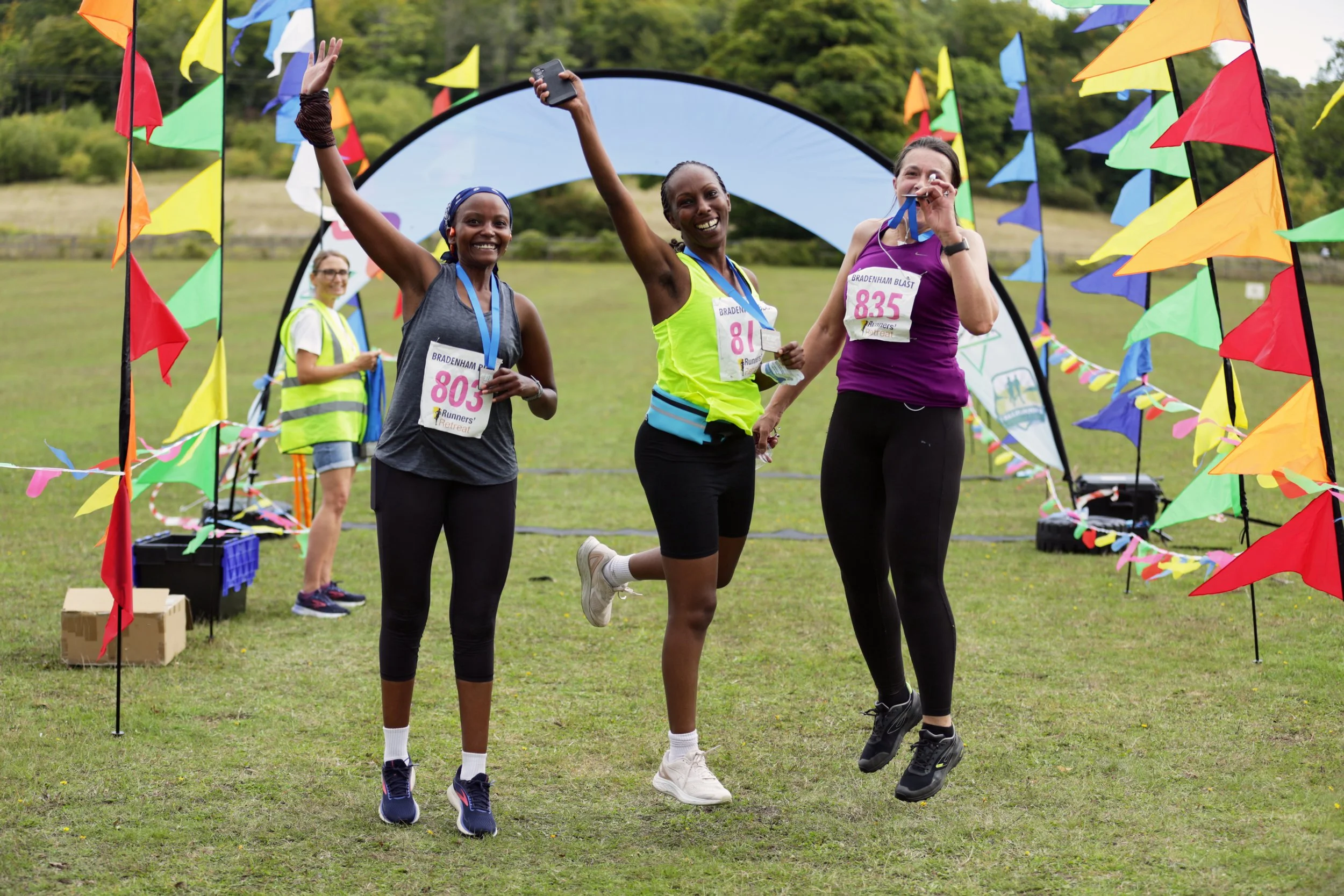 Three women celebrating at a race finish line, with flags and a backdrop of green trees, one is jumping, and the others are holding medals and smiling.