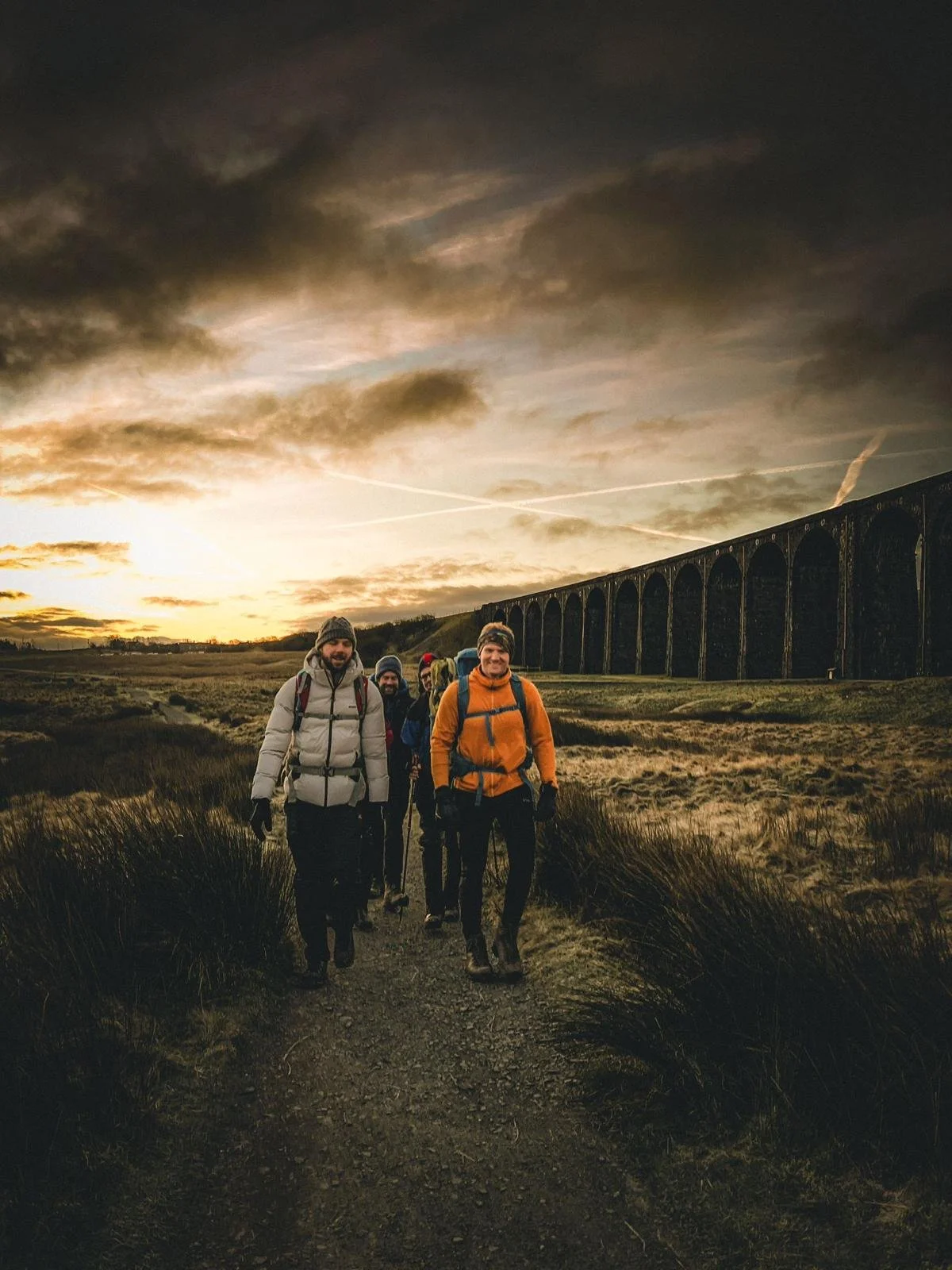 A group of five hikers walking along a trail at sunset with a viaduct in the background.