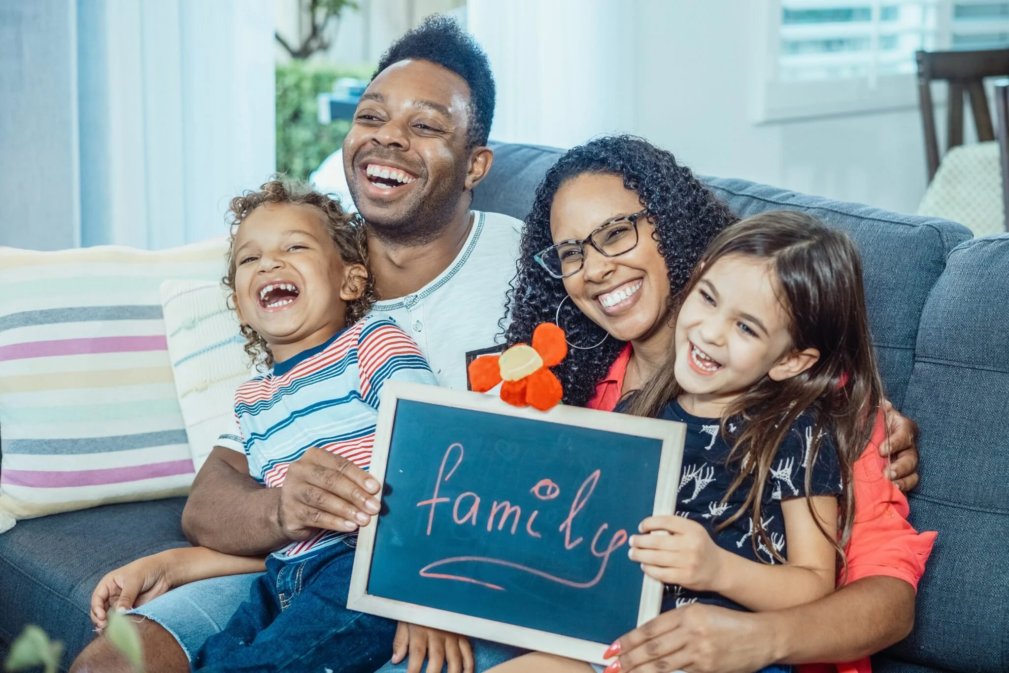 Family of four sitting on a couch, smiling and holding a chalkboard that says 'family' with a flower decoration.