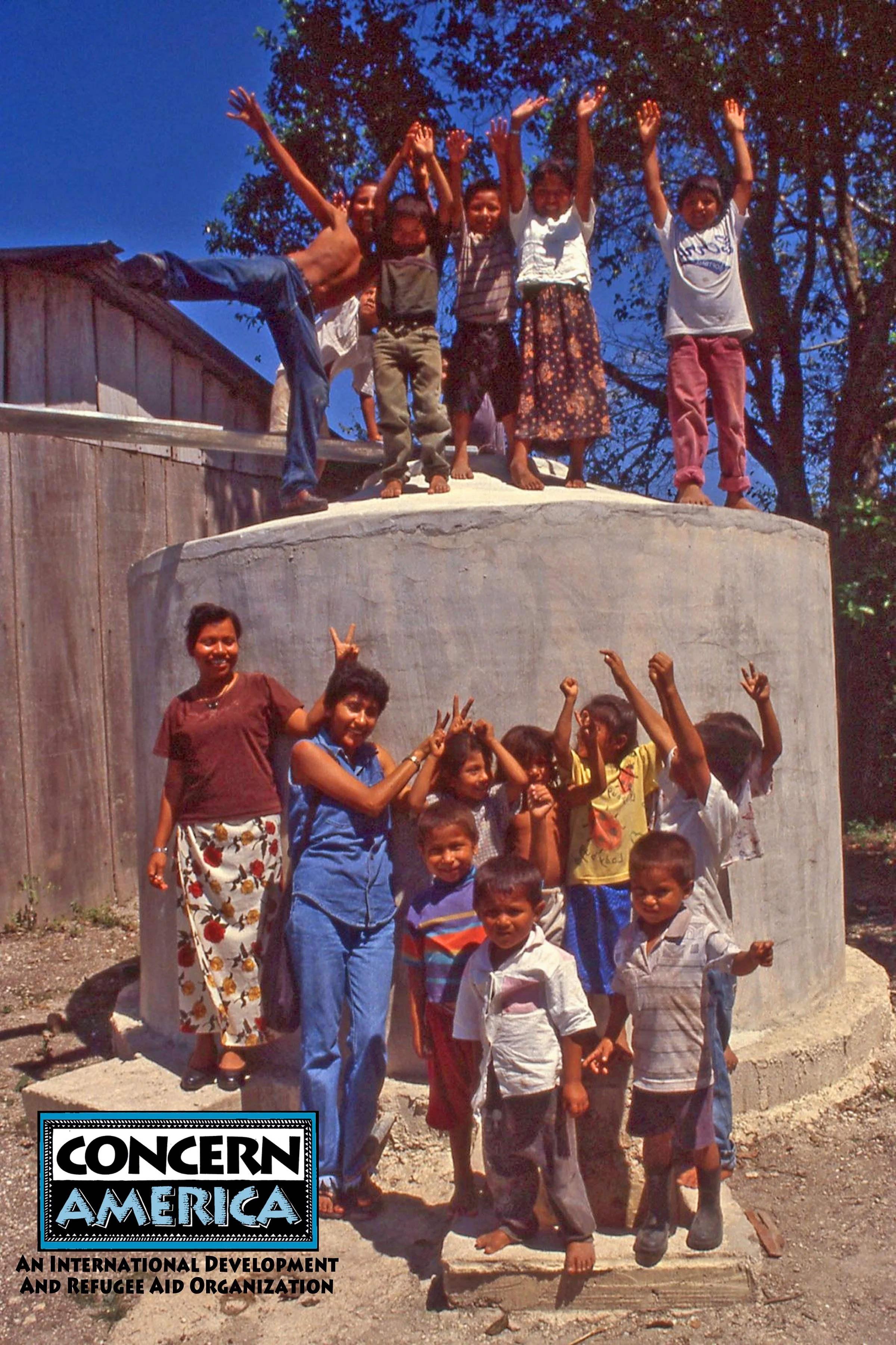 Children and adults smiling and making playful gestures around a large, cylindrical concrete structure outdoors. Some are standing on top of it, and others are standing in front. The scene appears sunny with a clear blue sky and trees in the background. The image has a logo of Concern America, an international development and refugee aid organization, in the bottom left corner.