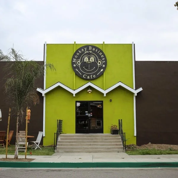 Front view of a bright green building with a monkey logo and sign that reads 'Monkey Business Cafe,' featuring a cartoon monkey face, stairs leading to the entrance, and outdoor seating including chairs and a table.
