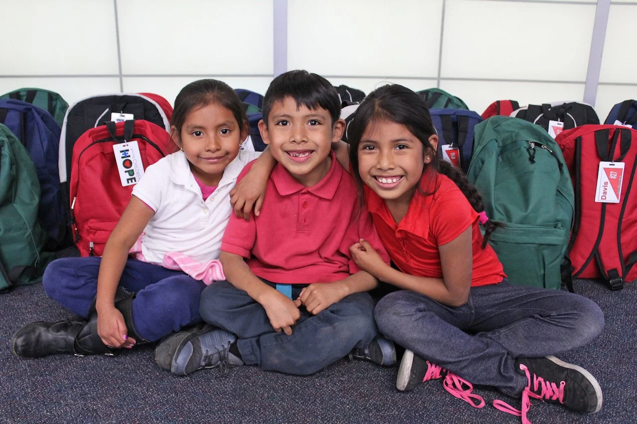 Three school children sitting on the floor with backpacks in the background, smiling at the camera.