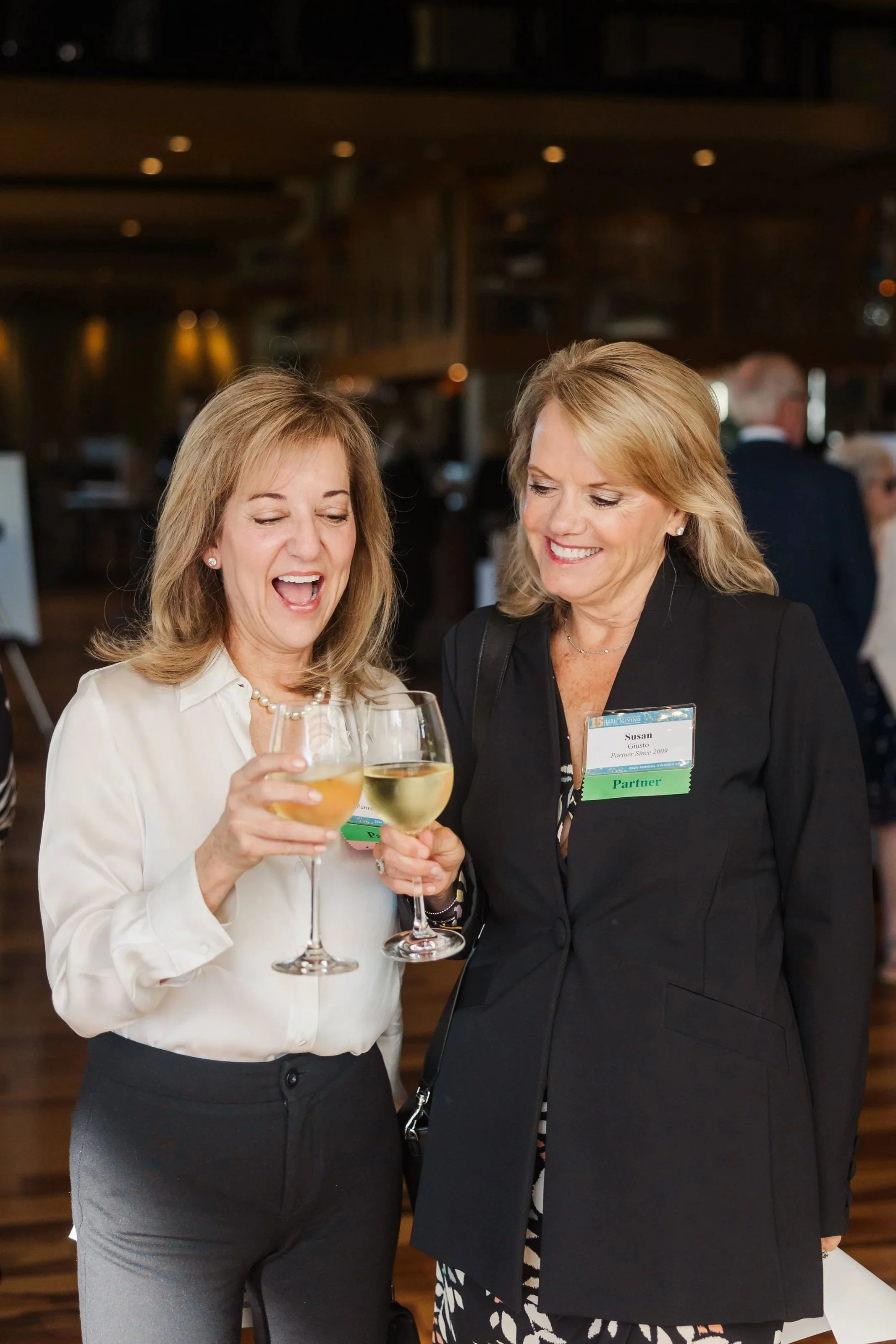 Two women smiling and hugging at an outdoor event, wearing name tags, with one wearing a black sleeveless top and the other a black and white striped blazer.