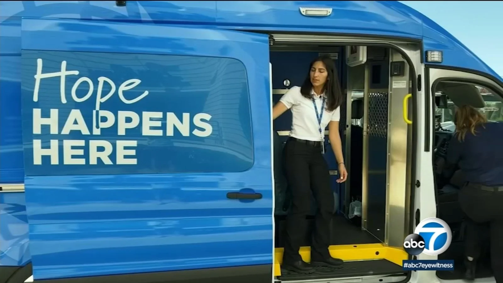 A blue mobile unit with the words 'Hope Happens Here' written on the side. Two women are present, one standing at the entrance and the other sitting inside the vehicle. The scene includes the ABC7 logo and hashtag #abc7eyewitness.
