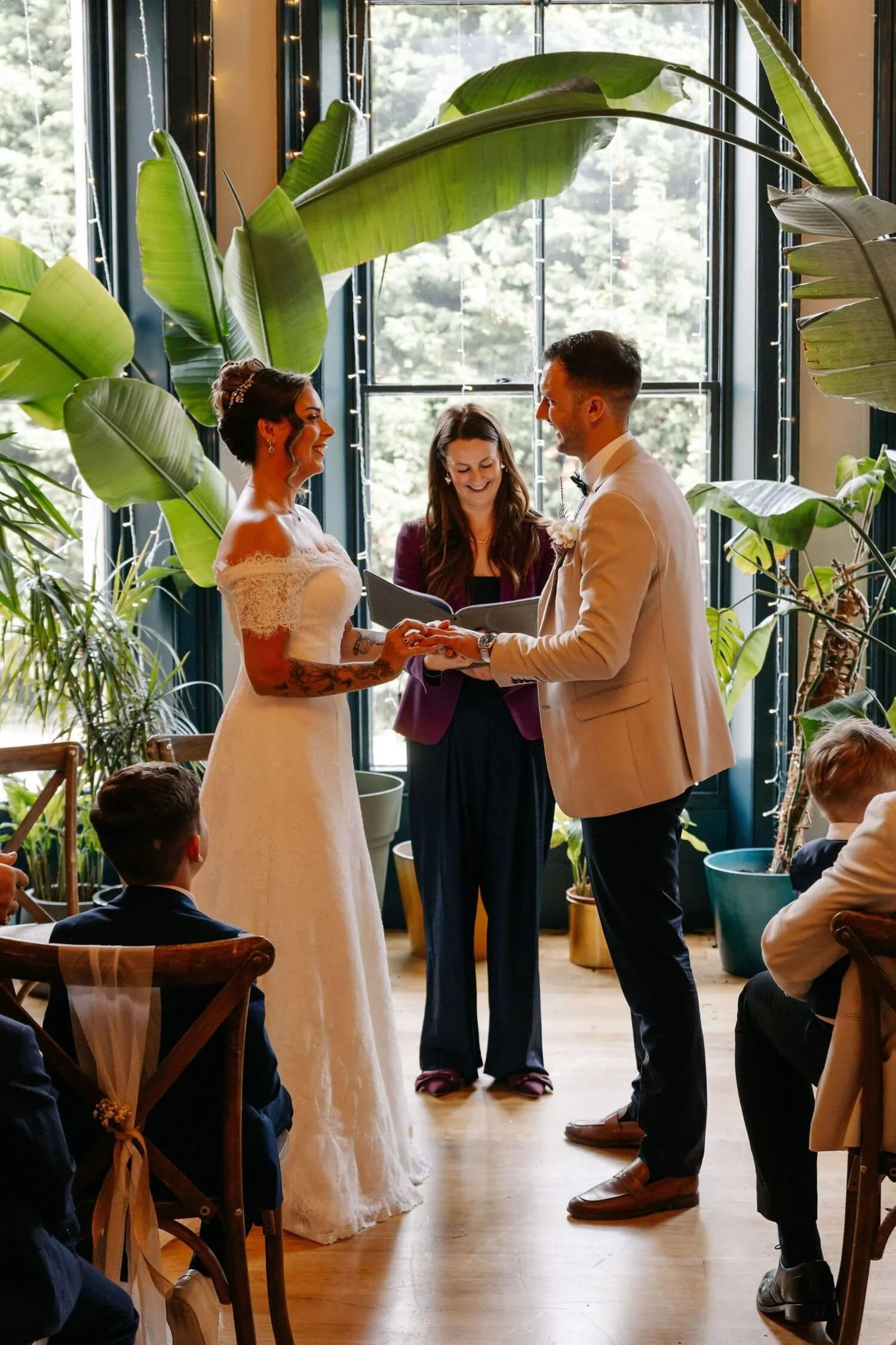 Celebrant ceremony at The Faversham in Leeds. Bride and Groom stand with Michelle Park celebrant in a ceremony room with green plants and a large window. 