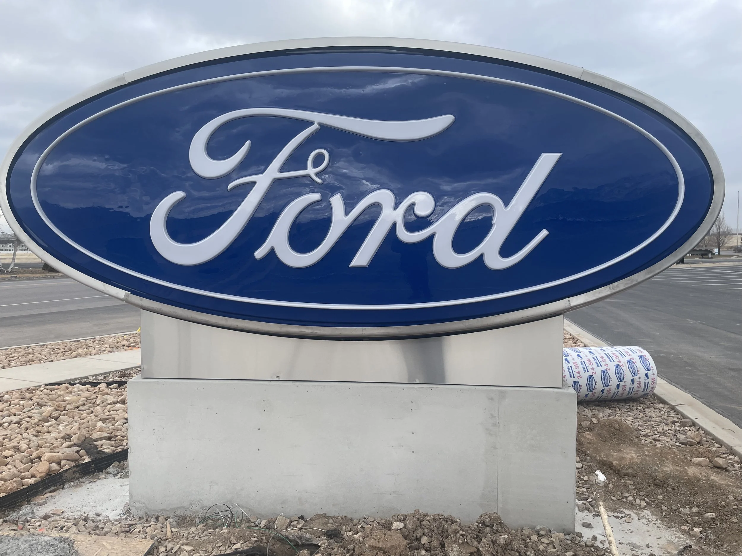 Close-up of a concrete base supporting a large Ford sign at an outdoor location with dirt, small rocks, a tree, and a road in the background.