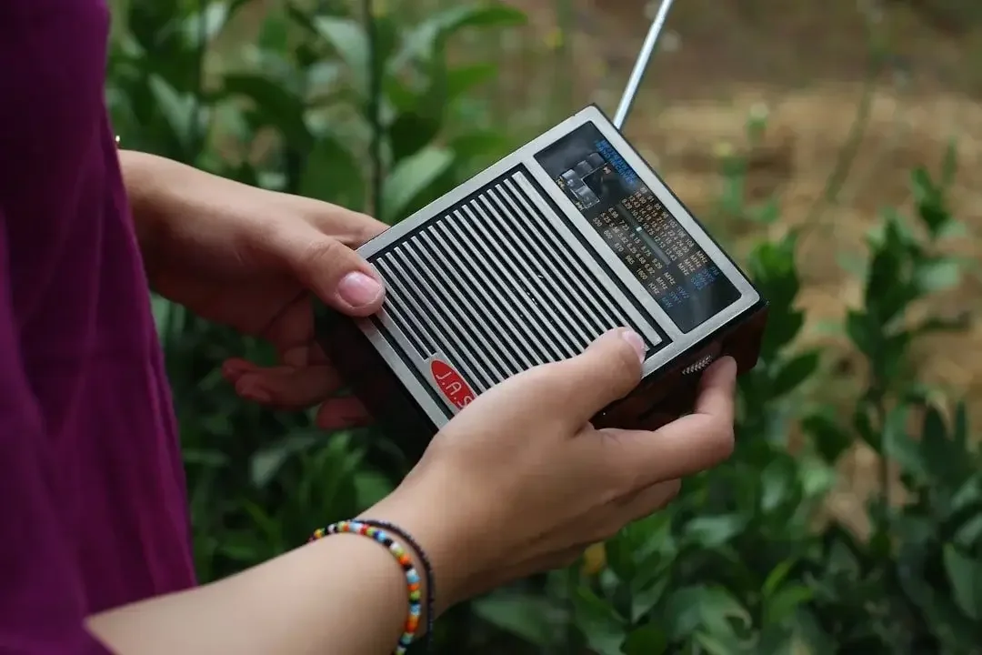 A person holding a small black radio with a digital display in a garden with green foliage.