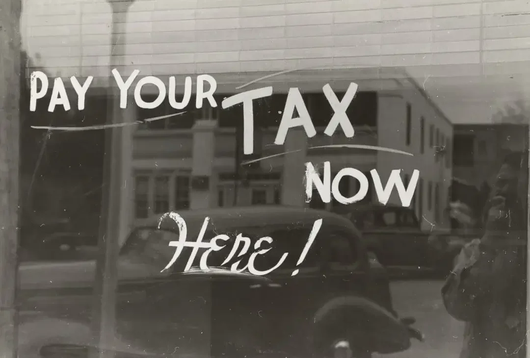 A reflection of a city street with buildings and cars on a window with the text 'Pay Your Tax Now Here!'.