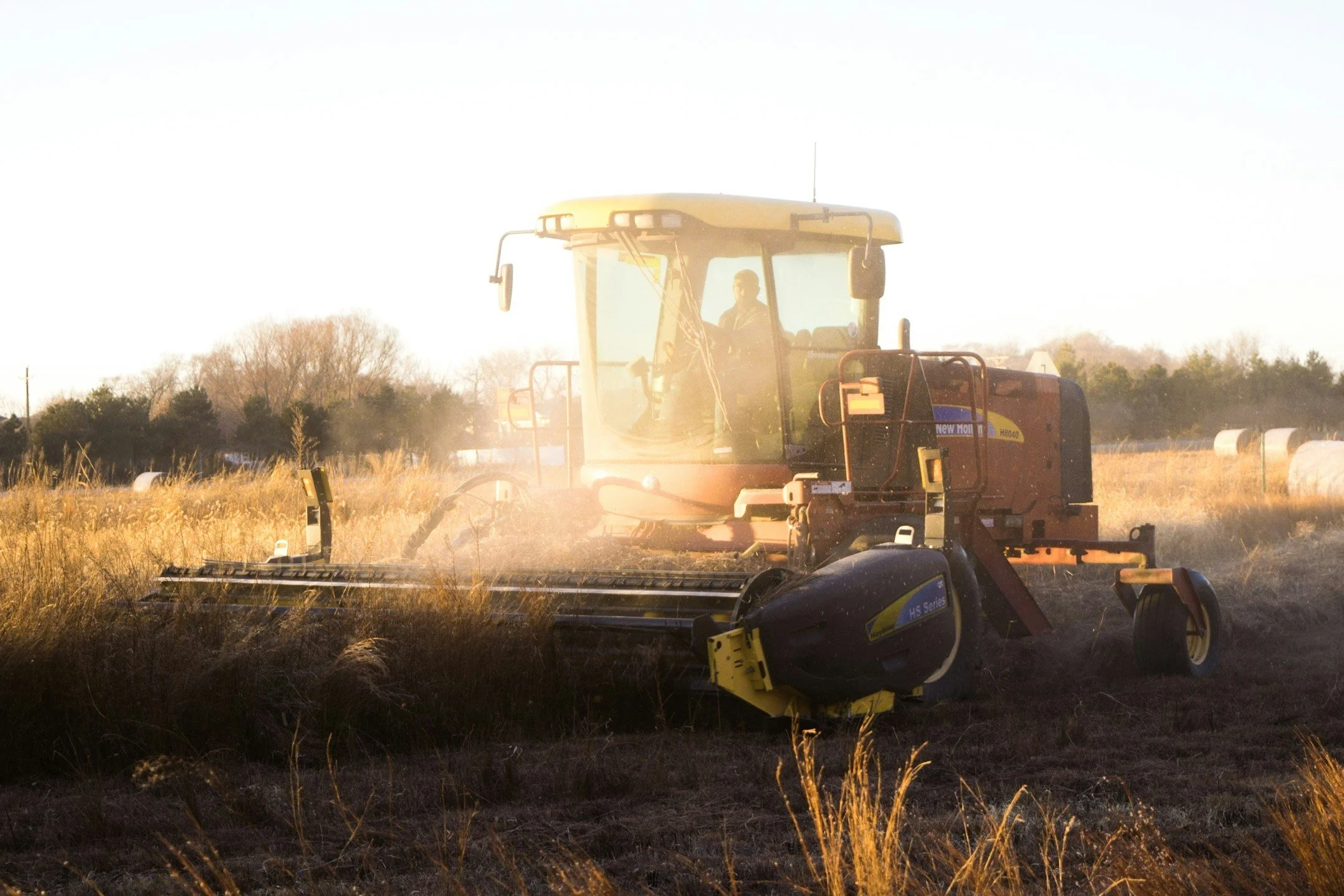 A person operating a red and black tractor in a field during sunset, with hay bales in the background.
