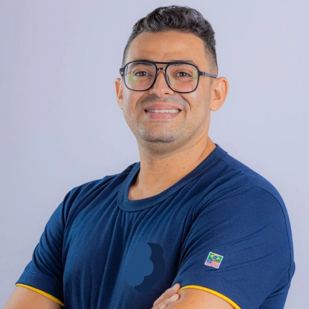 A man with glasses and short dark hair smiling, wearing a navy blue T-shirt with small yellow trim on the sleeves and a small Brazilian flag patch on the sleeve, posing with arms crossed against a light gray background.