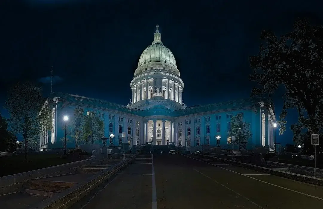 Illuminated state capitol building at night with a large central dome, trees, and street lamps in the foreground.