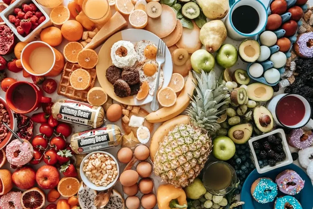 A colorful spread of breakfast foods including sliced oranges, bananas, kiwi, apples, pineapple, berries, eggs, breakfast meats, waffles, cupcakes, coffee, and juices.