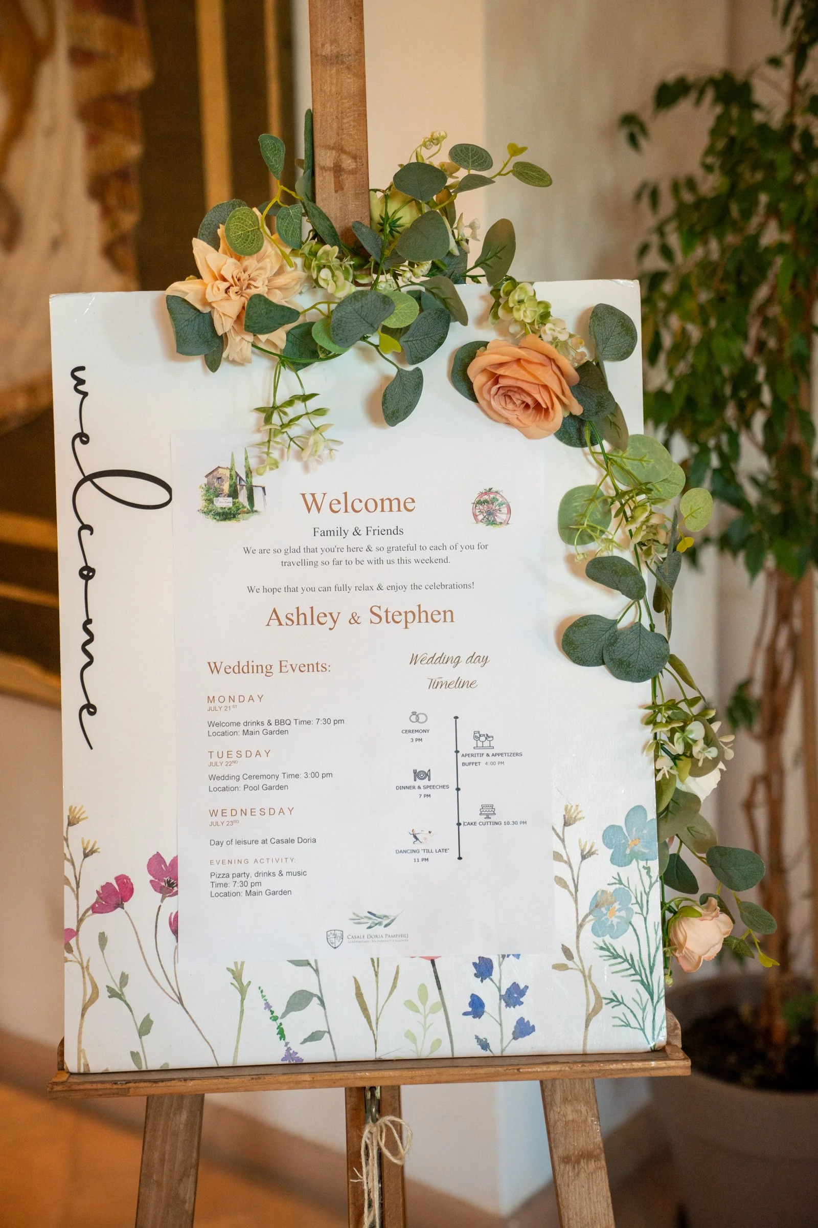 Wedding event schedule and welcome sign decorated with peach roses and green foliage on a wooden stand.