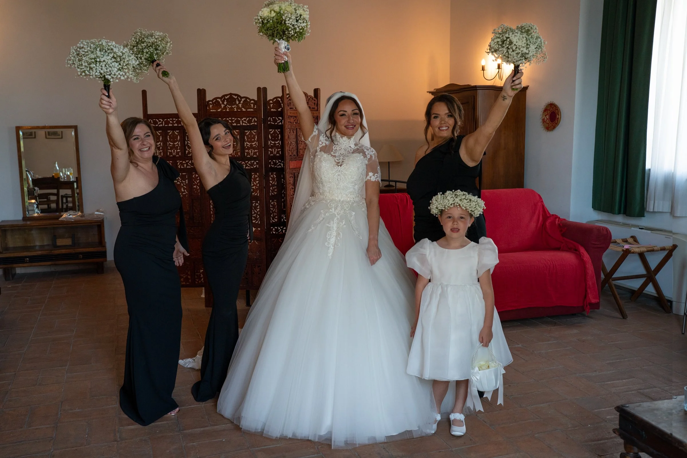 A bride in a white wedding gown and four women, three of whom are in black dresses and one in a white dress, holding floral bouquets and a flower crown, celebrating indoors with a red sofa and wooden furniture around.