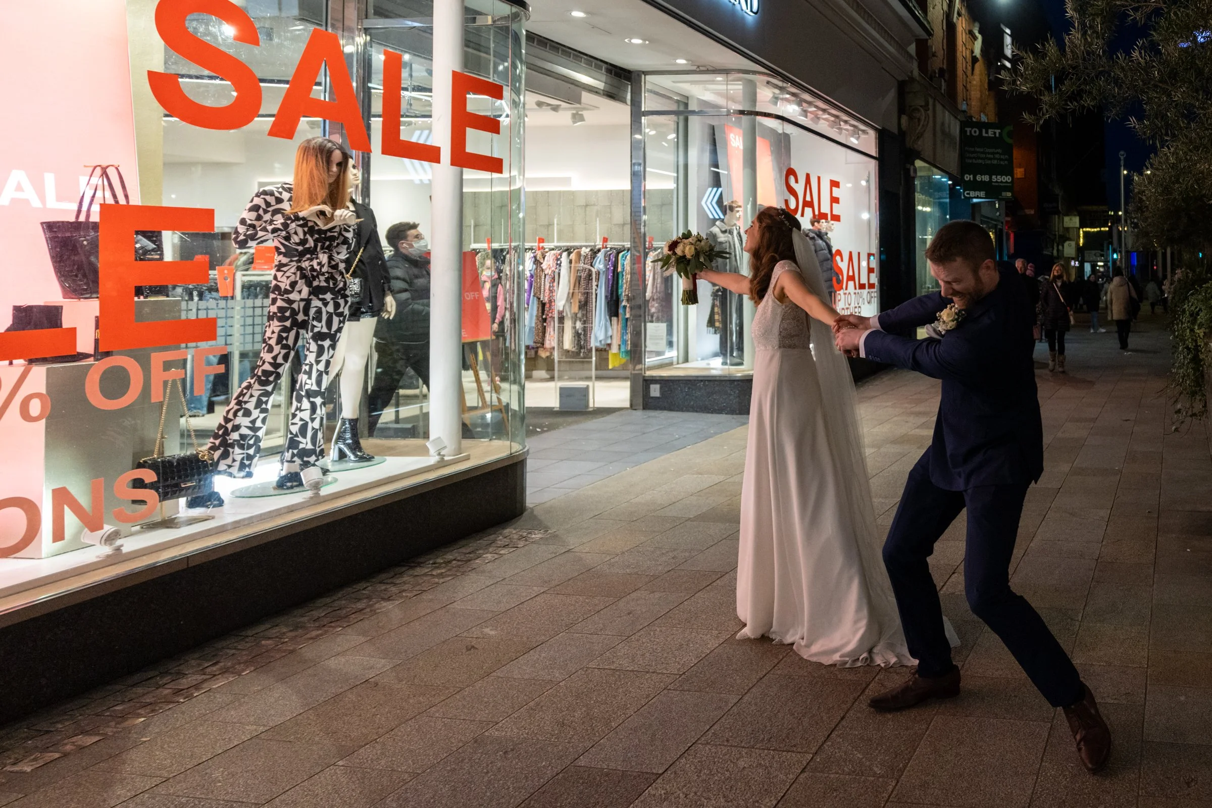 A woman in a wedding dress and a man in a suit are outside a clothing store with sale signs on the windows at night. The woman is holding a bouquet of flowers, and the man is pulling her by the hand in a playful manner.
