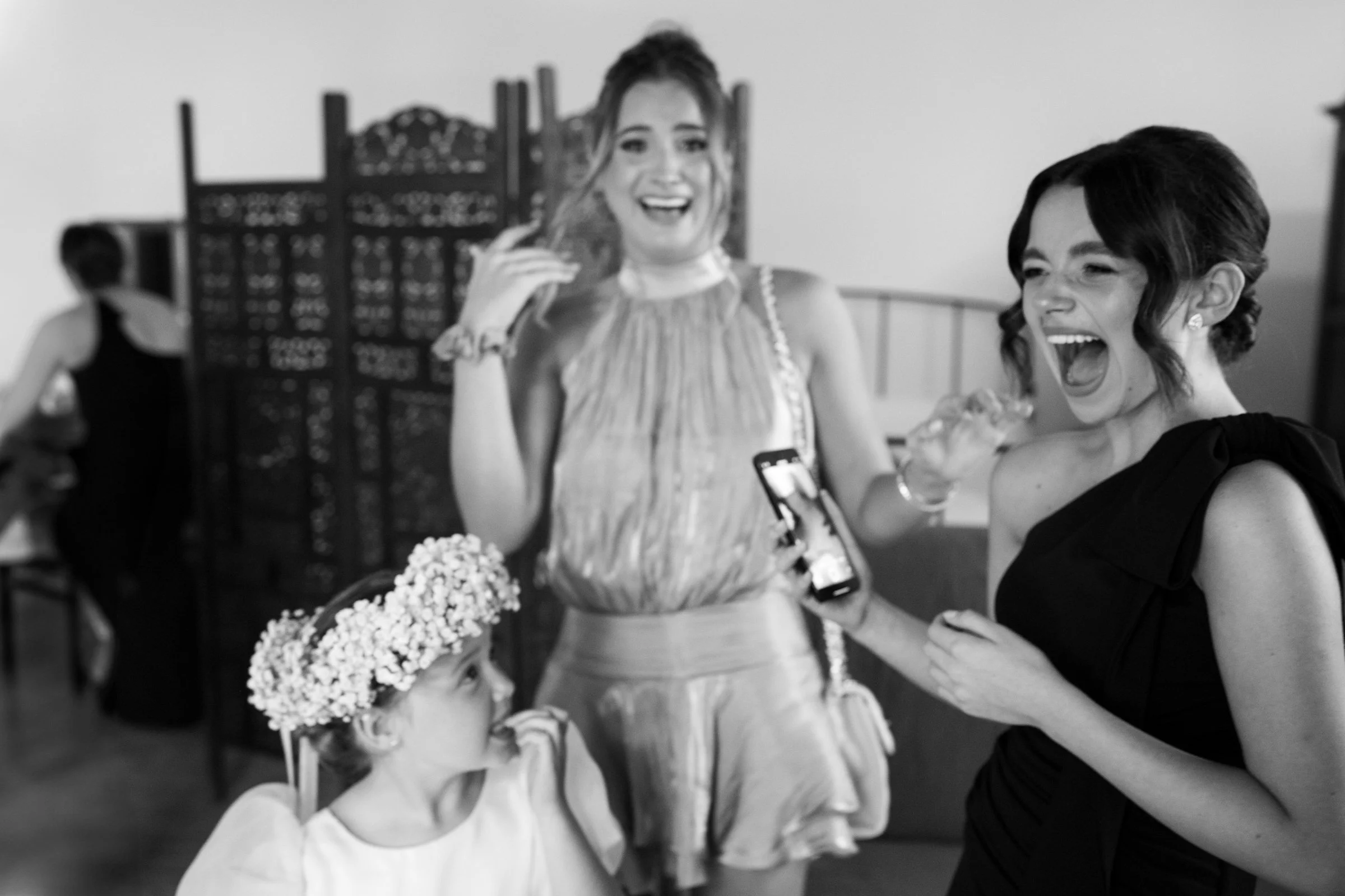 Three women and a young girl engaging in joyful conversation, with the women laughing and one woman holding a smartphone. The girl wears a floral crown and looks up at the women in a lively social setting.