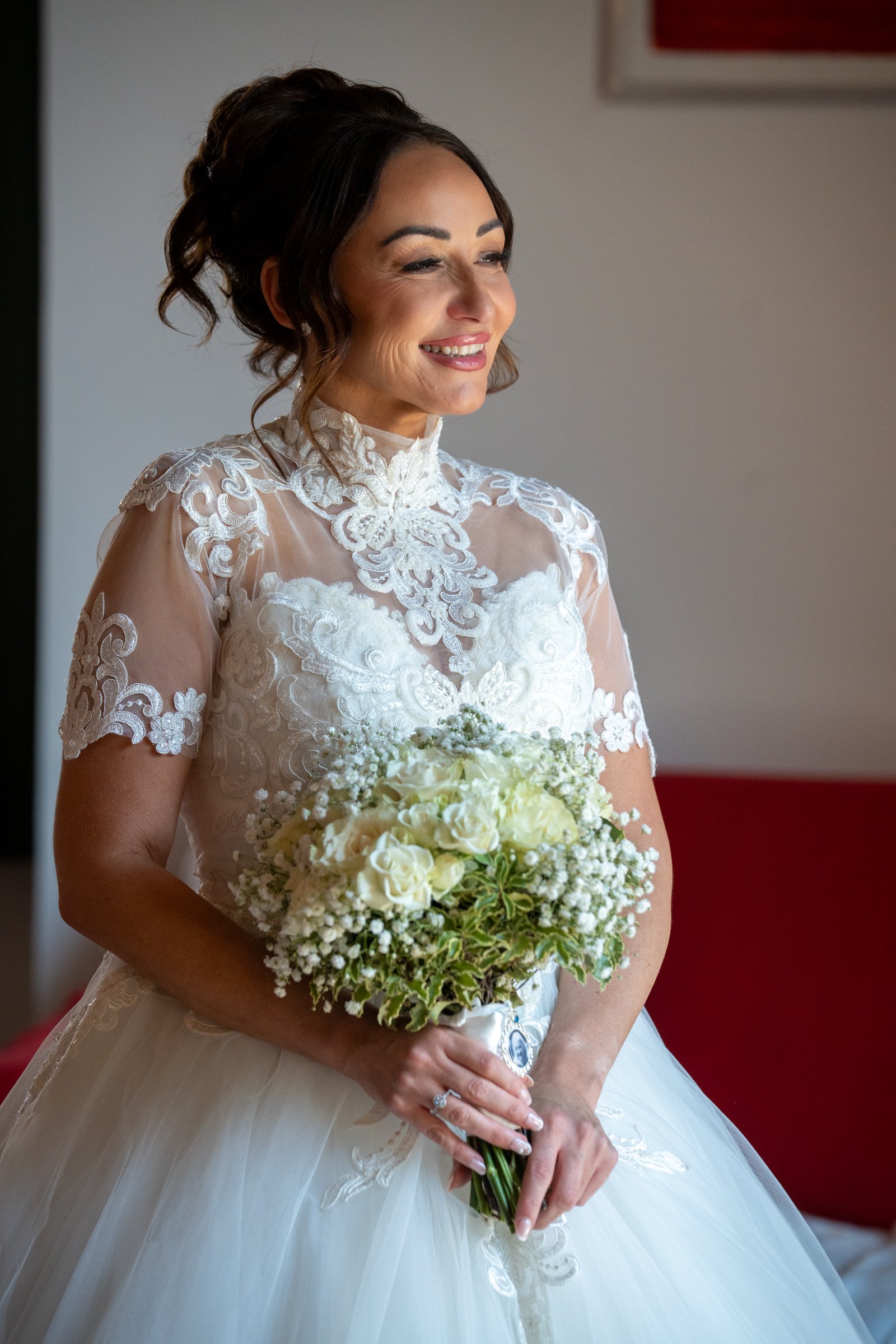 Bride with dark hair in an elegant lace wedding dress holding a bouquet of white roses and baby's breath, smiling and looking to the side.
