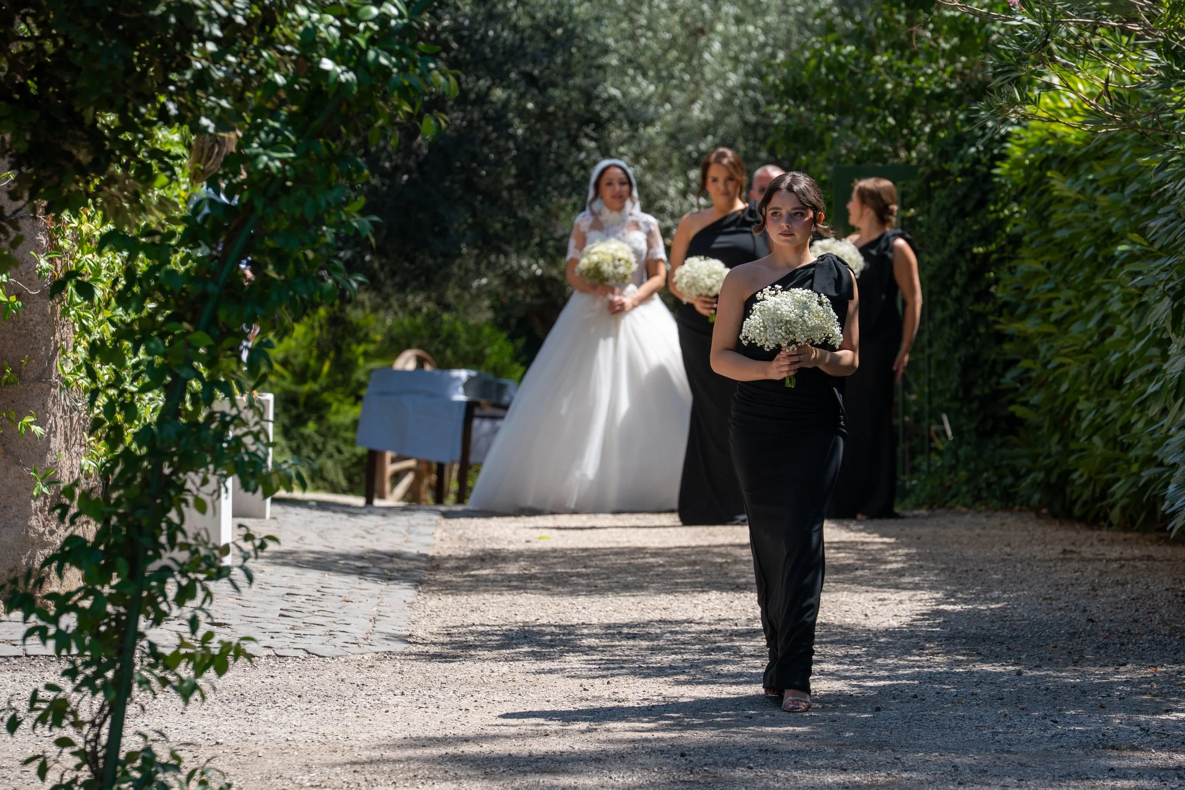 A bride in a white wedding dress and veil is standing with her bridesmaids dressed in black, holding white bouquets, outdoors surrounded by greenery.