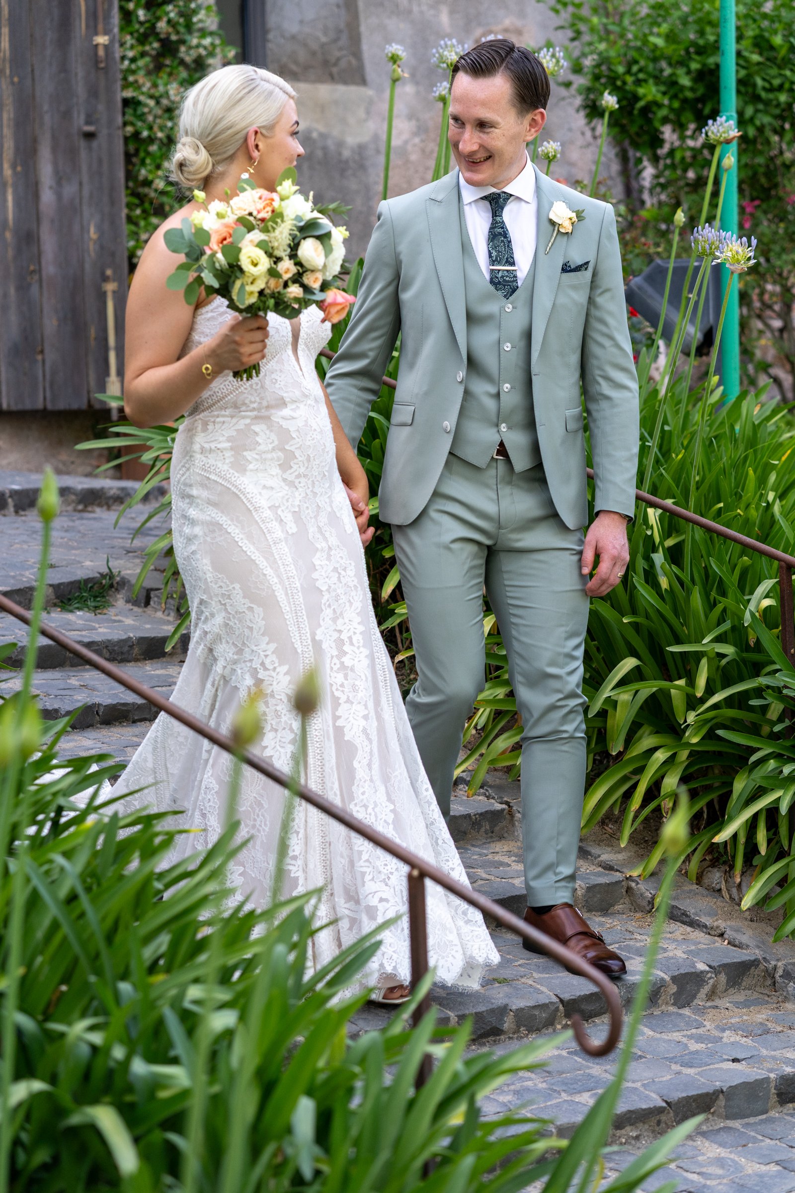 A bride and groom holding hands and smiling at each other outdoors during a wedding ceremony. The bride wears a white lace wedding dress and holds a bouquet of flowers. The groom is dressed in a light gray suit with a white shirt and patterned tie.