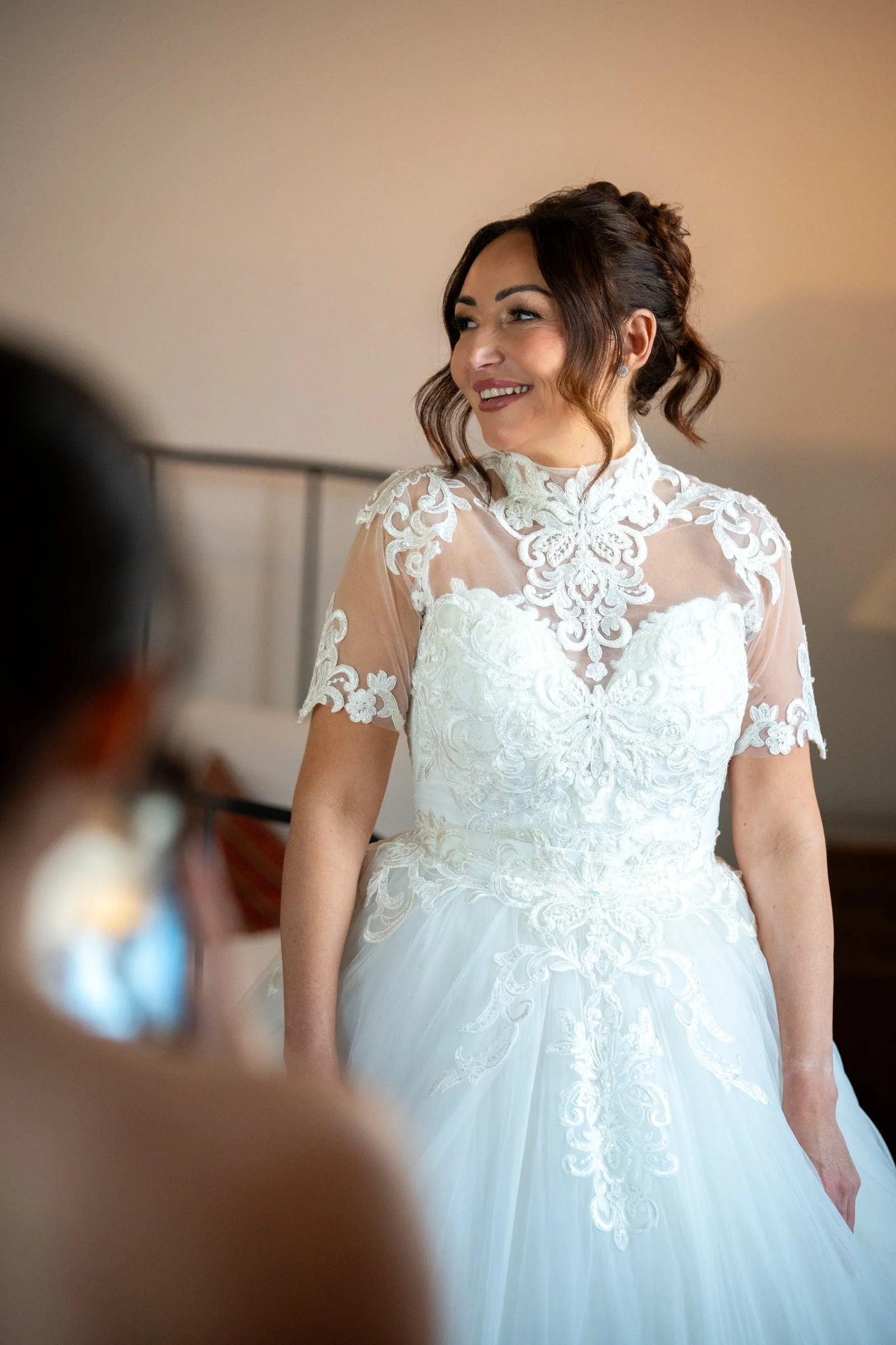 A woman in a white wedding dress with lace details, smiling and looking to her left, inside a room.