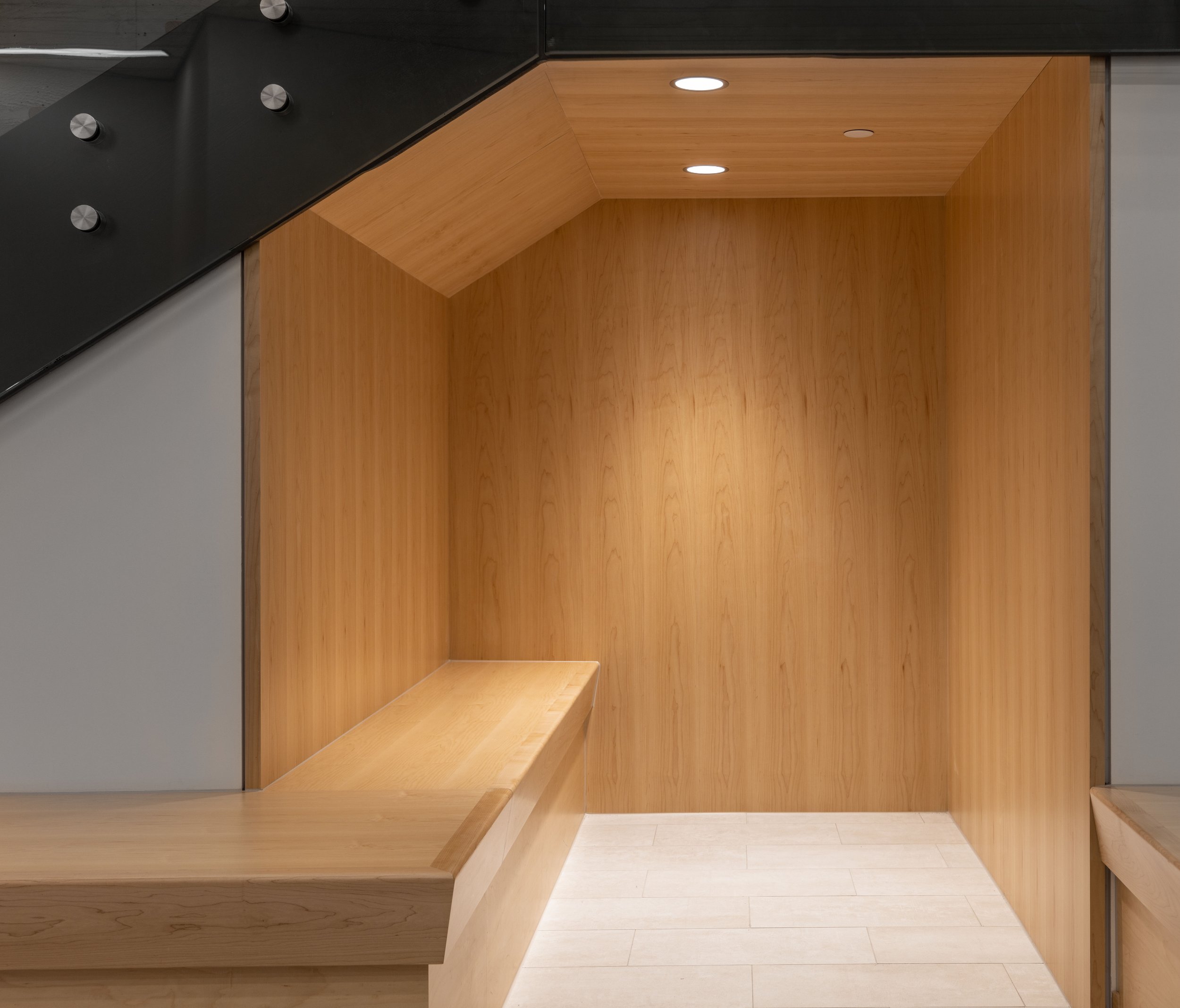 An open sitting area underneath a set of stairs. Wood paneling lines the space, two pot lights sit on the ceiling, and a large wooden bench wraps around the side of the stairs into the space.