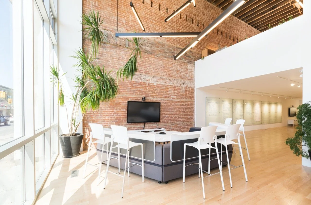 Open meeting space where a high table is attached to the back of a couch and has extra seating. A tv is mounted on the wall in front of the couch.