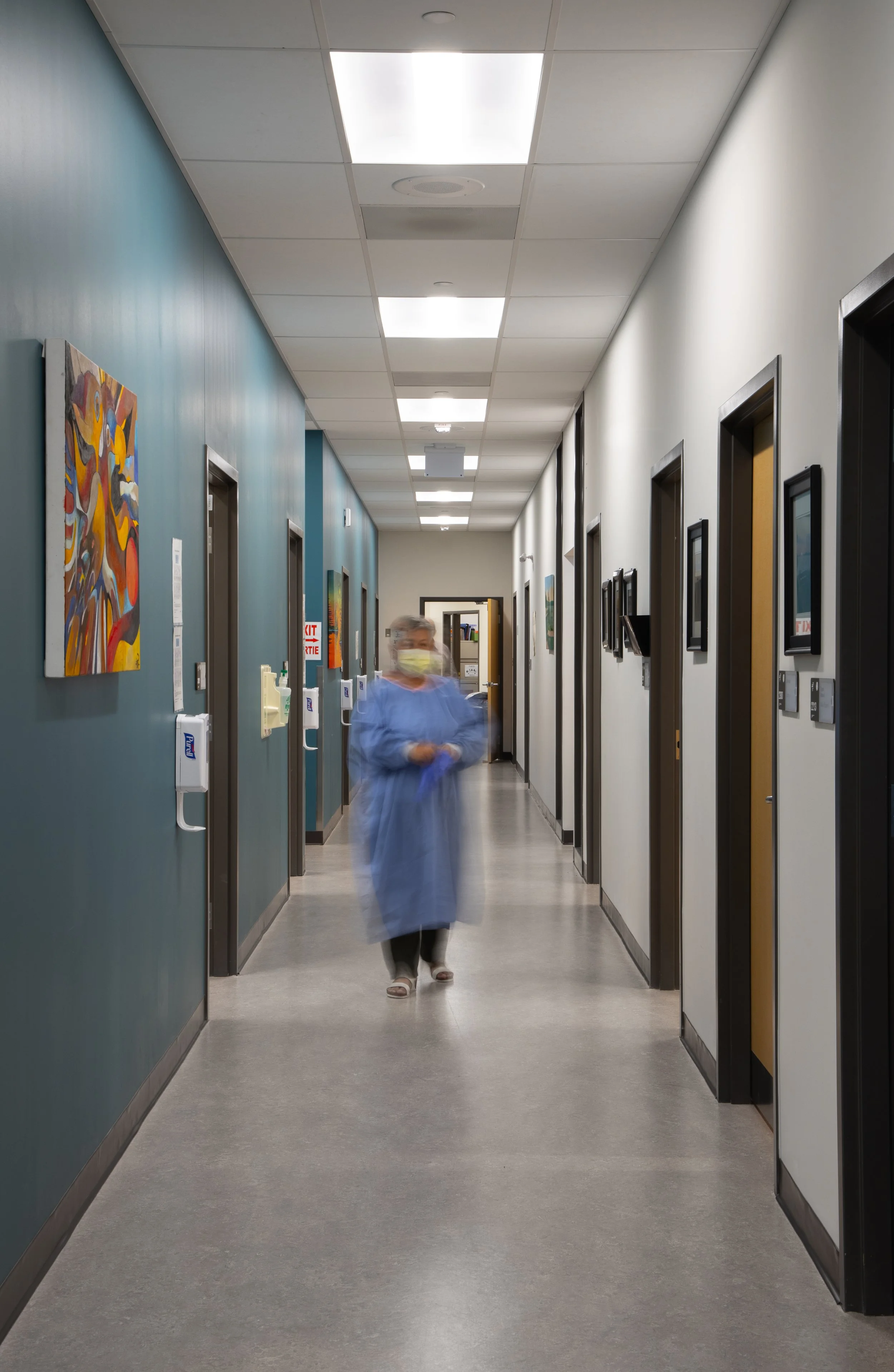 A medical professional, wearing a yellow mask and blue gown is blurred as they put on blue gloves in the middle of the hallway