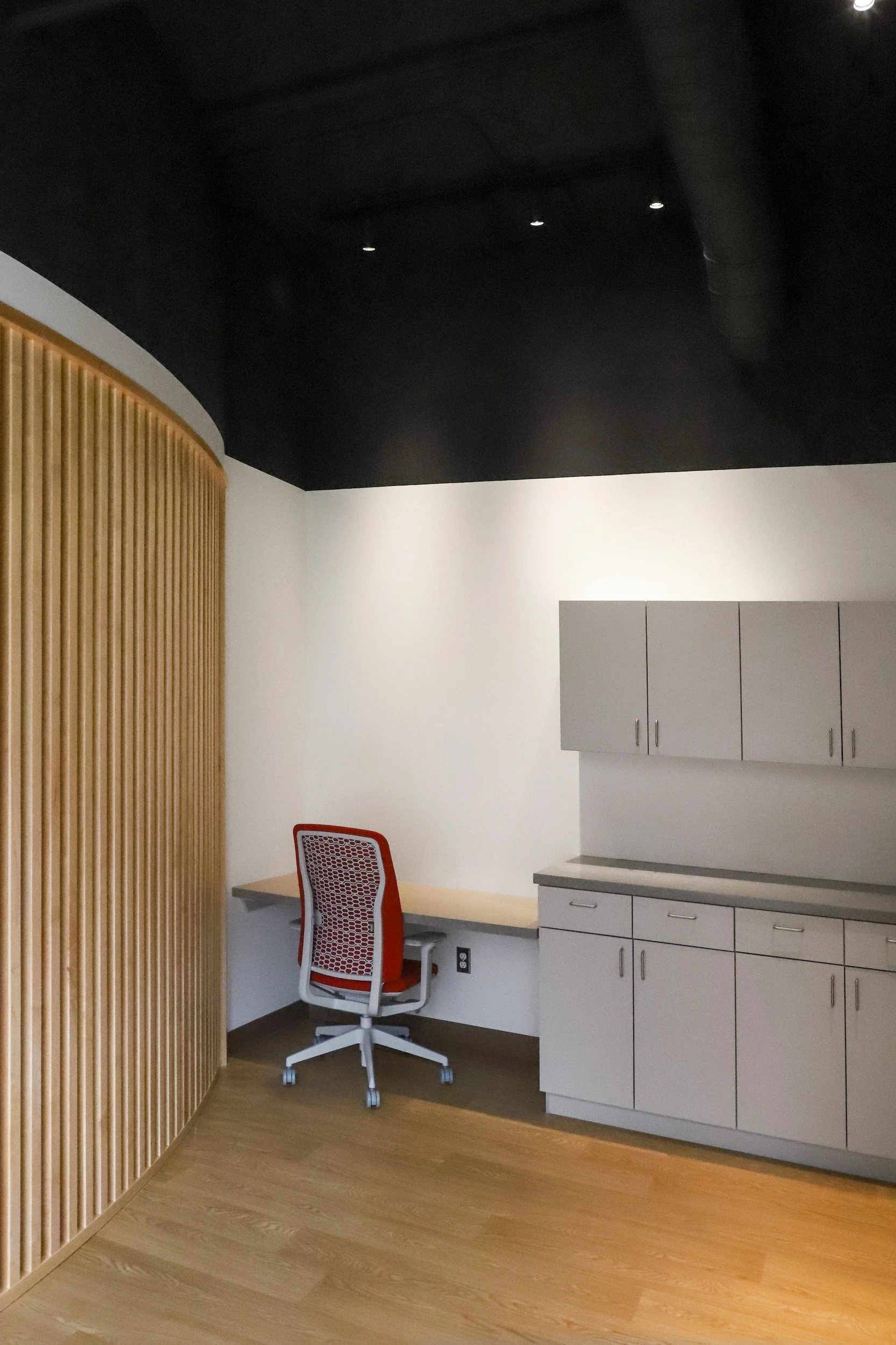 Modern office interior with a red ergonomic chair, built-in desk, gray cabinets, wood slat accent wall, and black ceiling