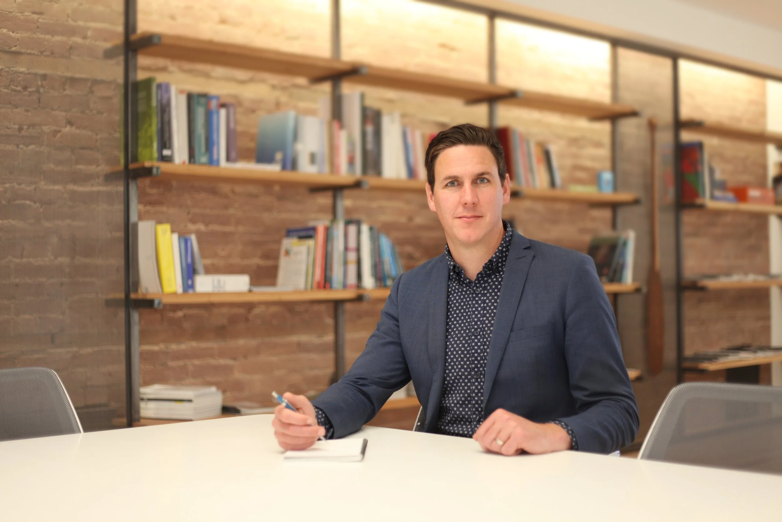 Michael sits resting both hands on the table with a pen in his left left, he sits smiling at the camera.