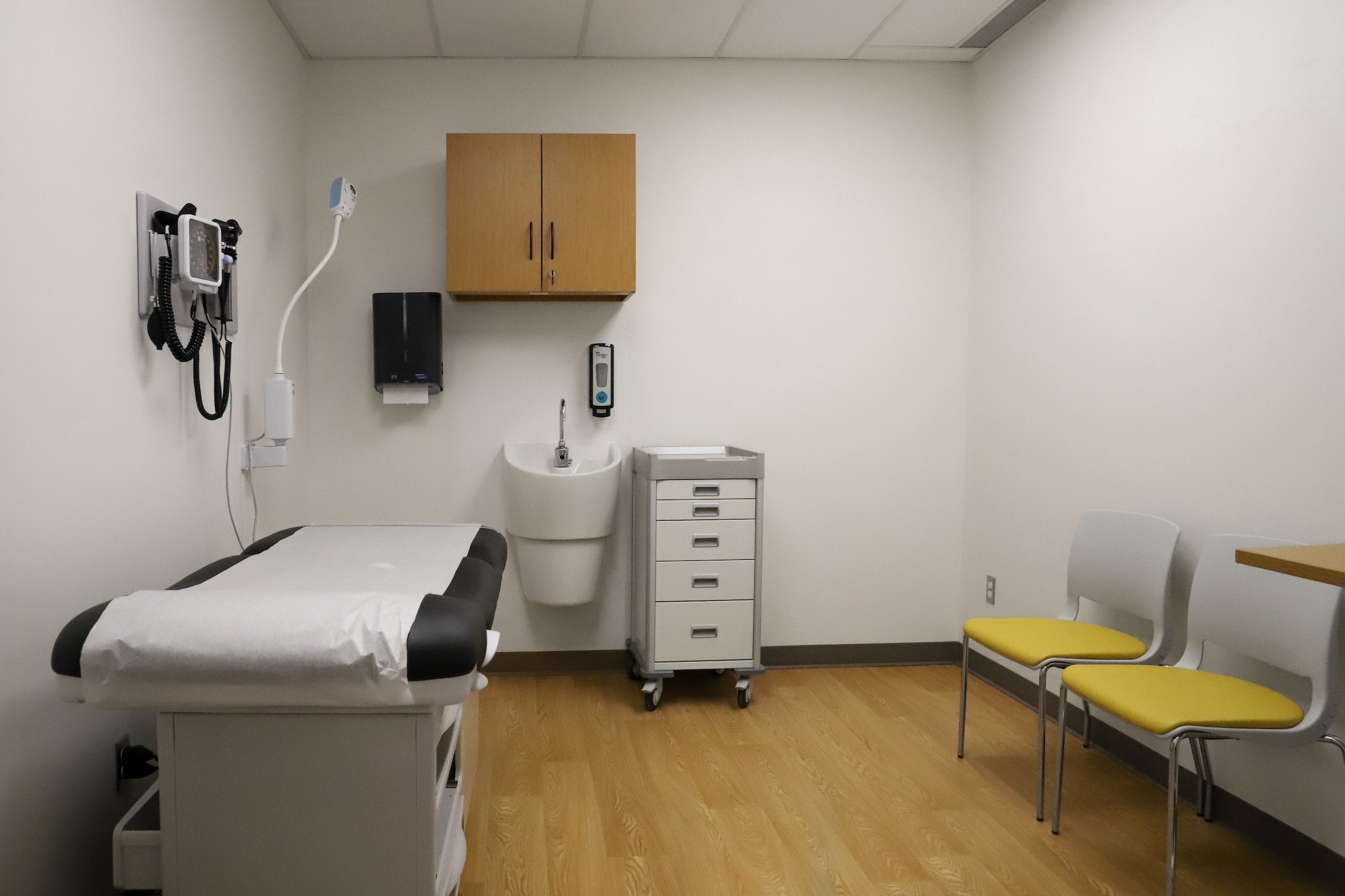 Empty medical examination room with exam bed, wall-mounted diagnostic equipment, sink, storage cabinet, rolling medical cart, and yellow chairs.