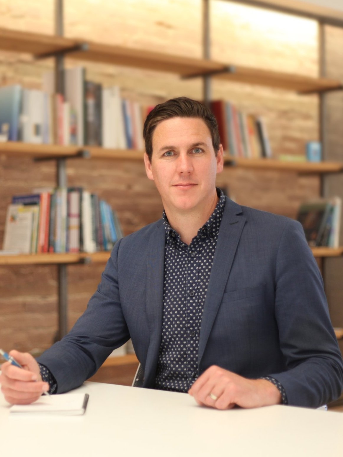 Michael sits resting both hands on the table with a pen in his left left, he sits smiling at the camera.