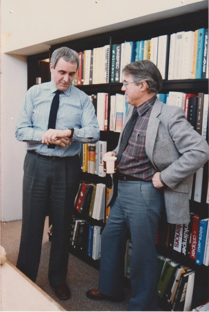 Bill Lett and Peter Smith stand in front of a bookcase having a conversation. Peter looks at his watch, Bill holds a coffee in his right hand.
