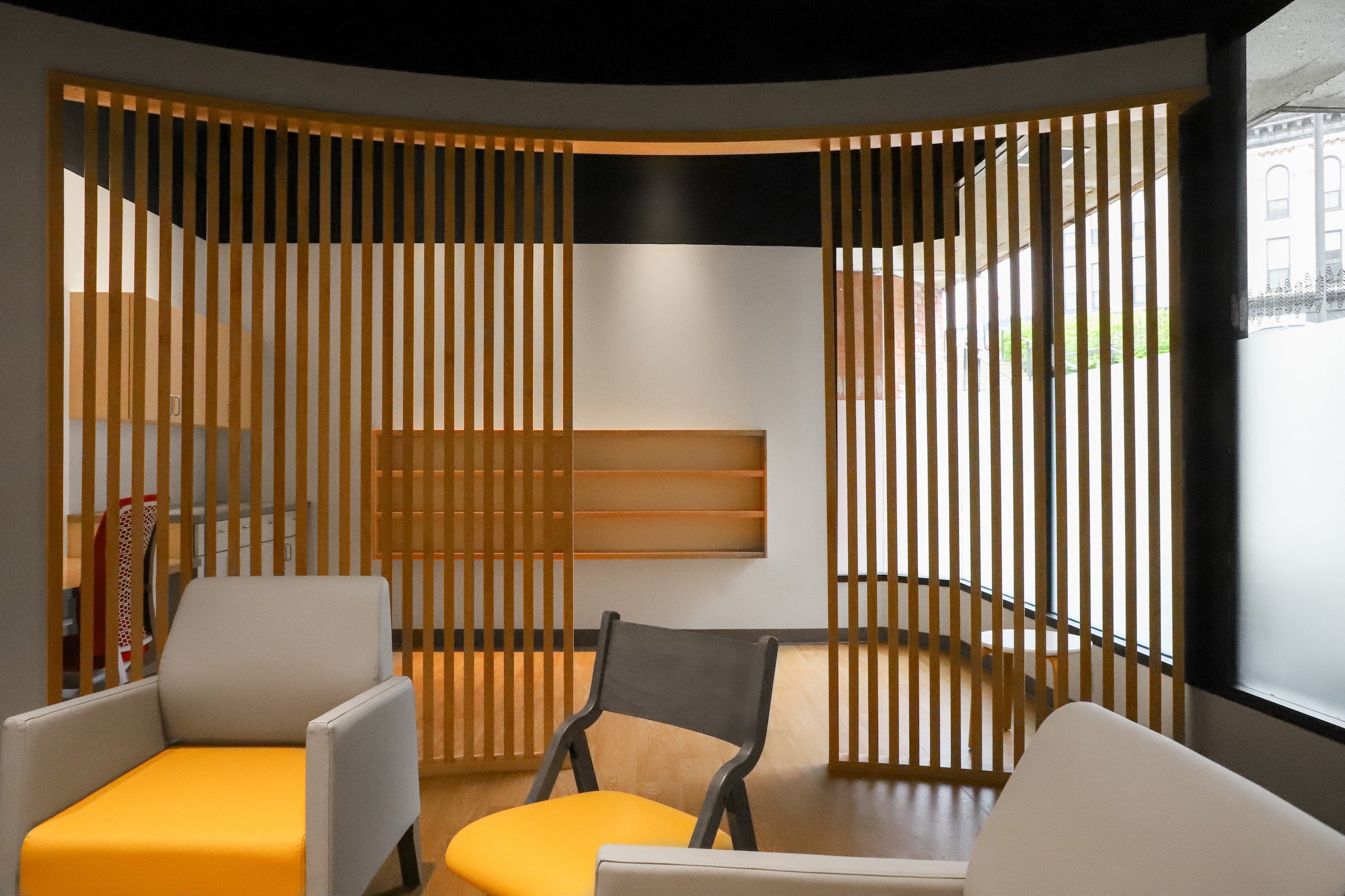 Modern lounge with yellow and gray chairs, wood slat partition, built-in shelving, and curved windows