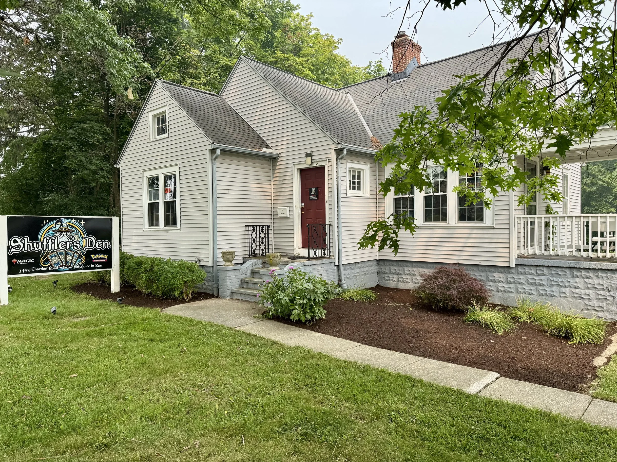A white house with a red door, multiple windows, and a sloped roof, surrounded by a garden with plants, trees, and a sidewalk, and a sign in the yard that says 'Shuffler's Den'.