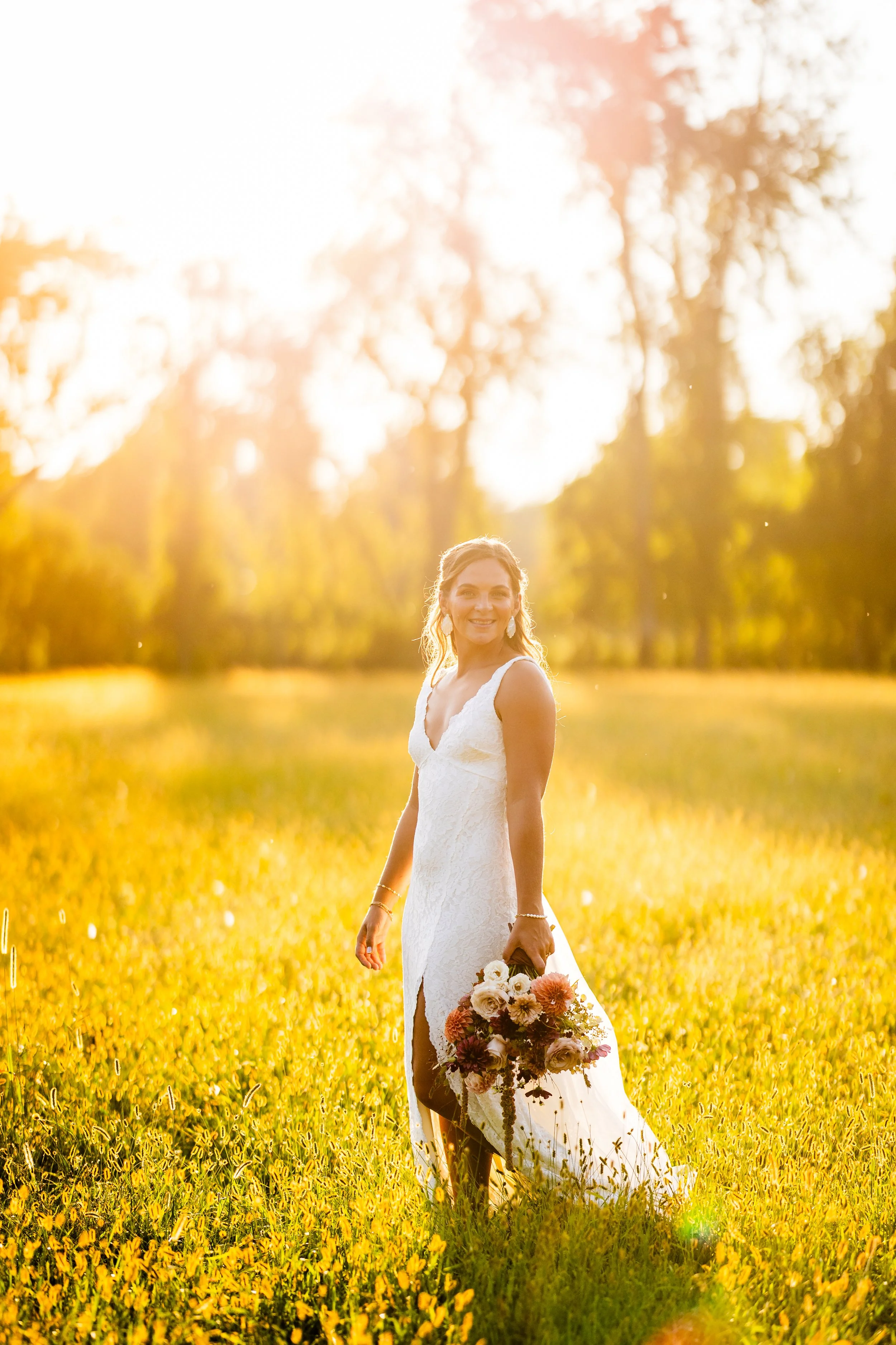 A woman in a white dress standing in a sunlit field holding a bouquet of flowers, with trees and sunlight in the background.