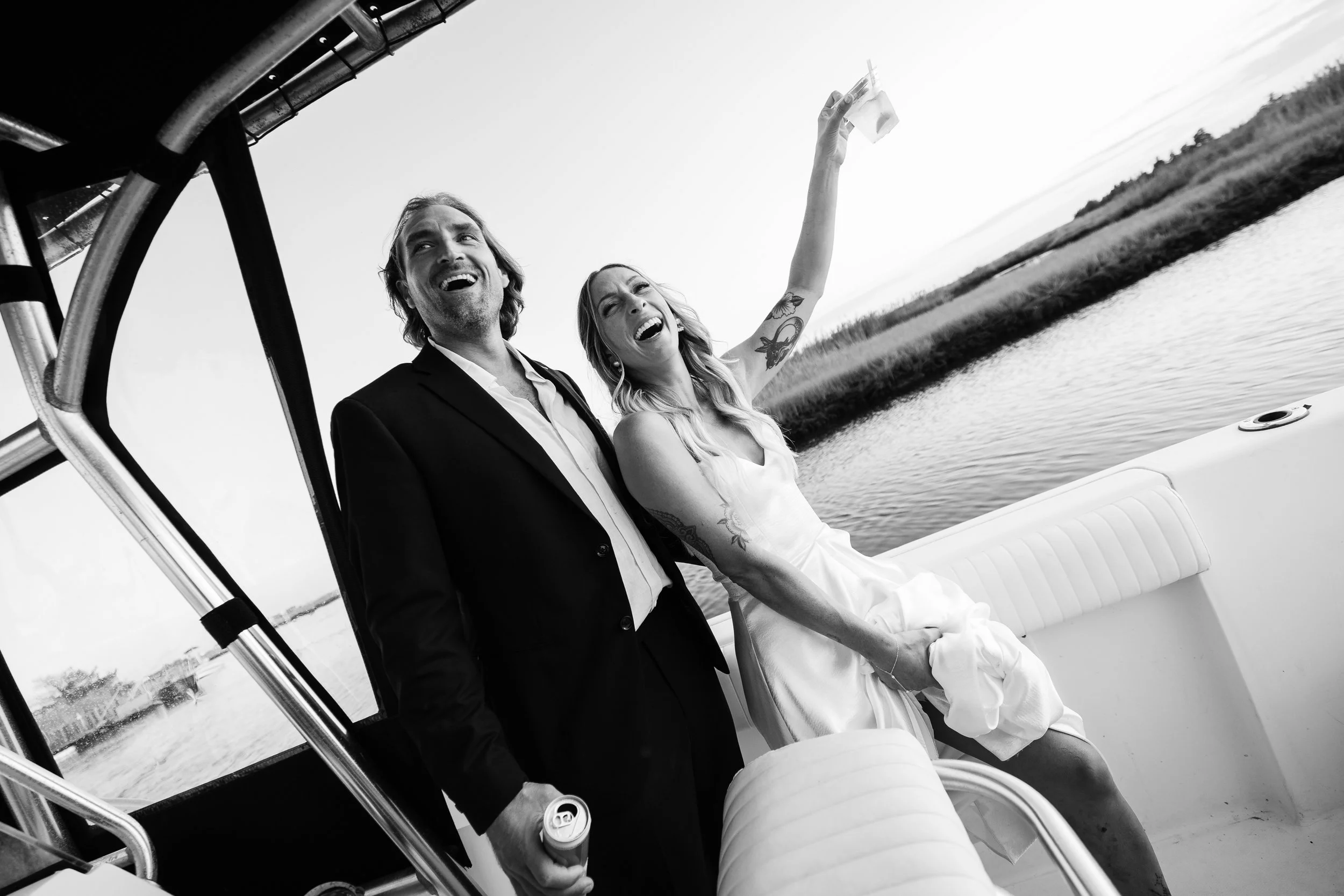 A man in a suit and a woman in a white dress are on a boat, laughing and celebrating, with the woman holding a drink in the air.