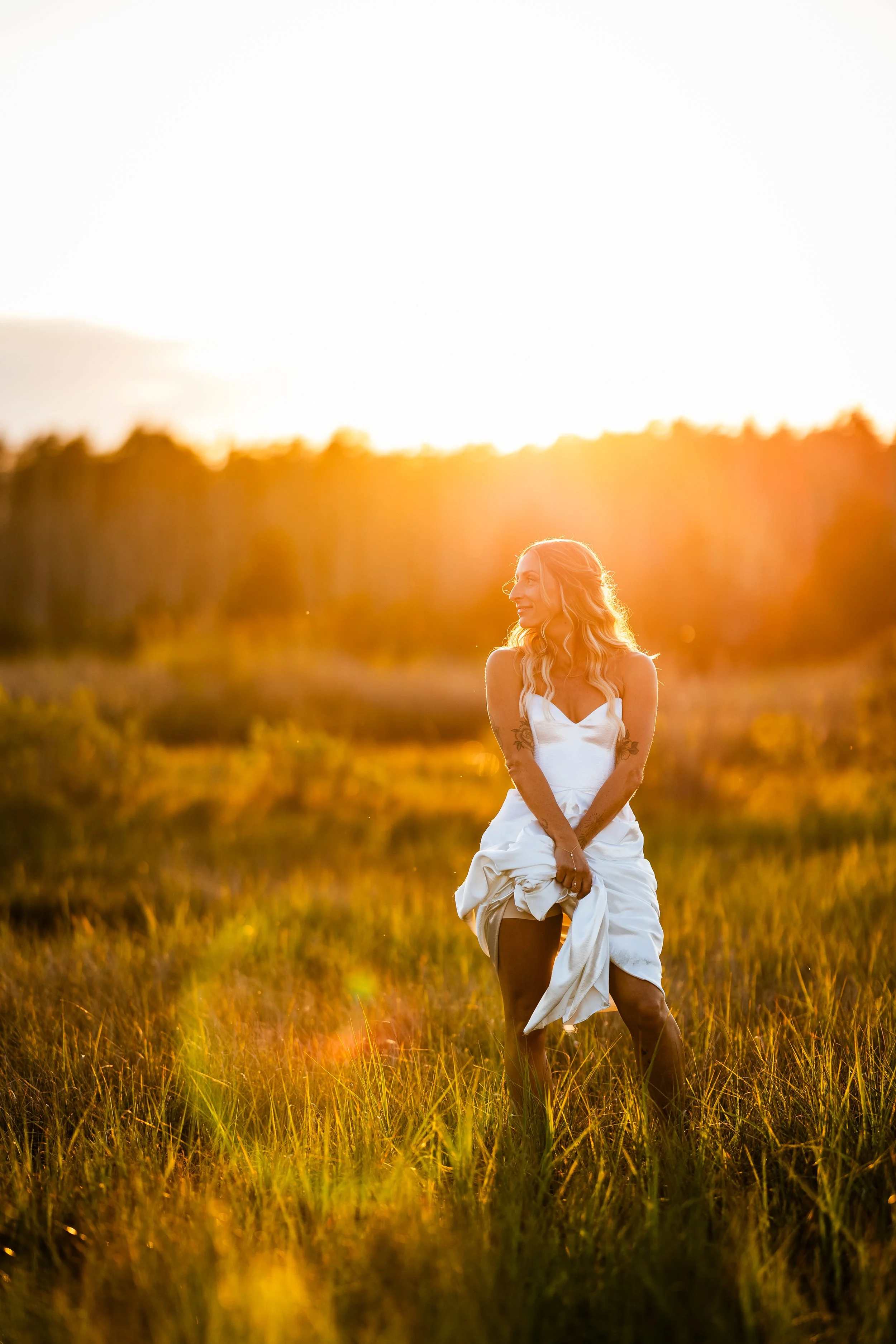 A woman in a white dress standing in a grassy field during sunset, smiling and looking to her left.